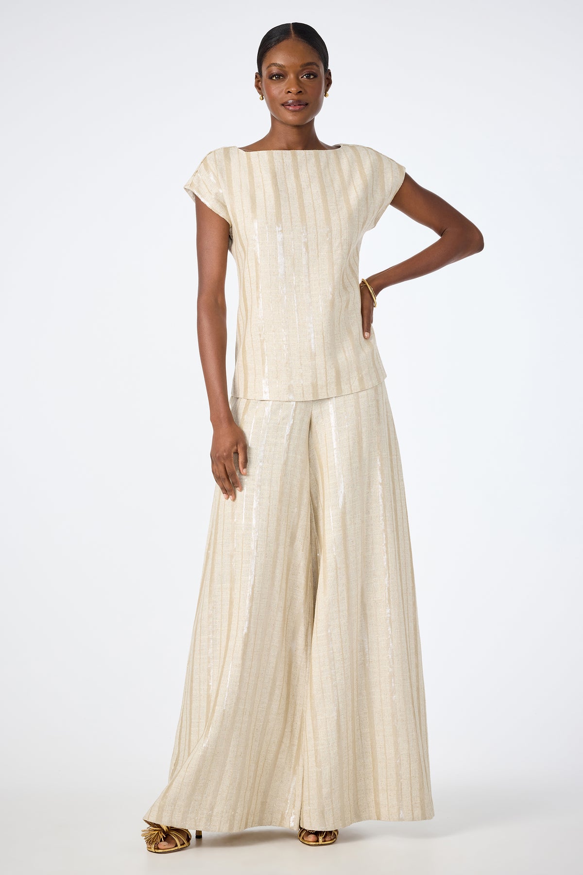 A woman stands confidently in the Breene Boatneck Top, a light gold, shimmery short-sleeve piece paired with wide-leg pants and gold heels. Her hair is pulled back as she poses against a plain white background.