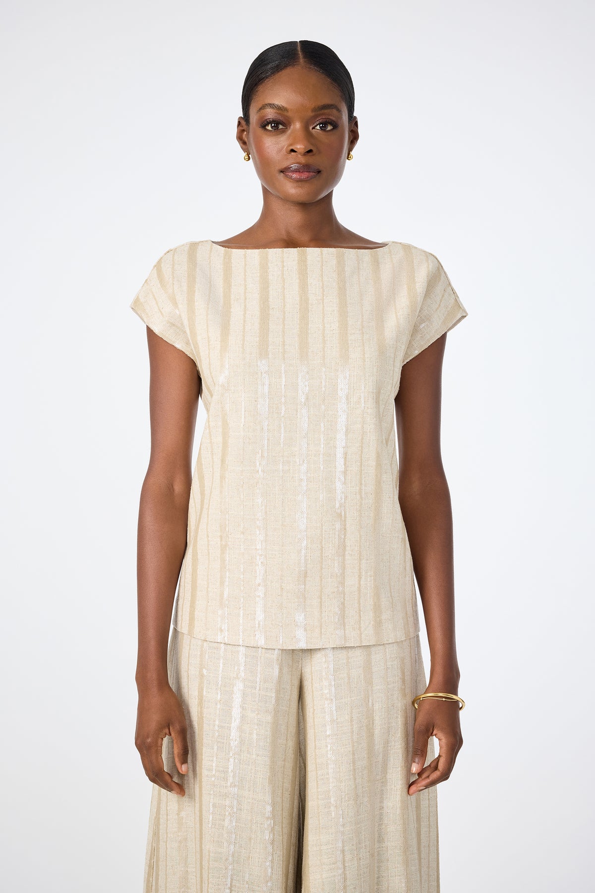A woman stands facing forward in the Breene Boatneck Top in Biarritz Beige, featuring short sleeves and stripes, paired with matching wide-leg pants. She has her hair pulled back and accessorizes with small gold earrings and a bracelet against a white background.