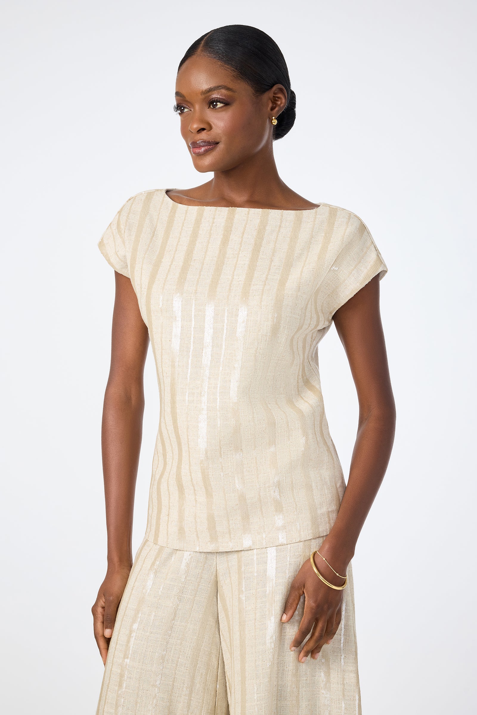 A woman wears the Breene Boatneck Top—a light beige, short-sleeve, striped blouse—paired with matching wide-leg pants, standing against a plain white background and smiling gently as she looks to the side.