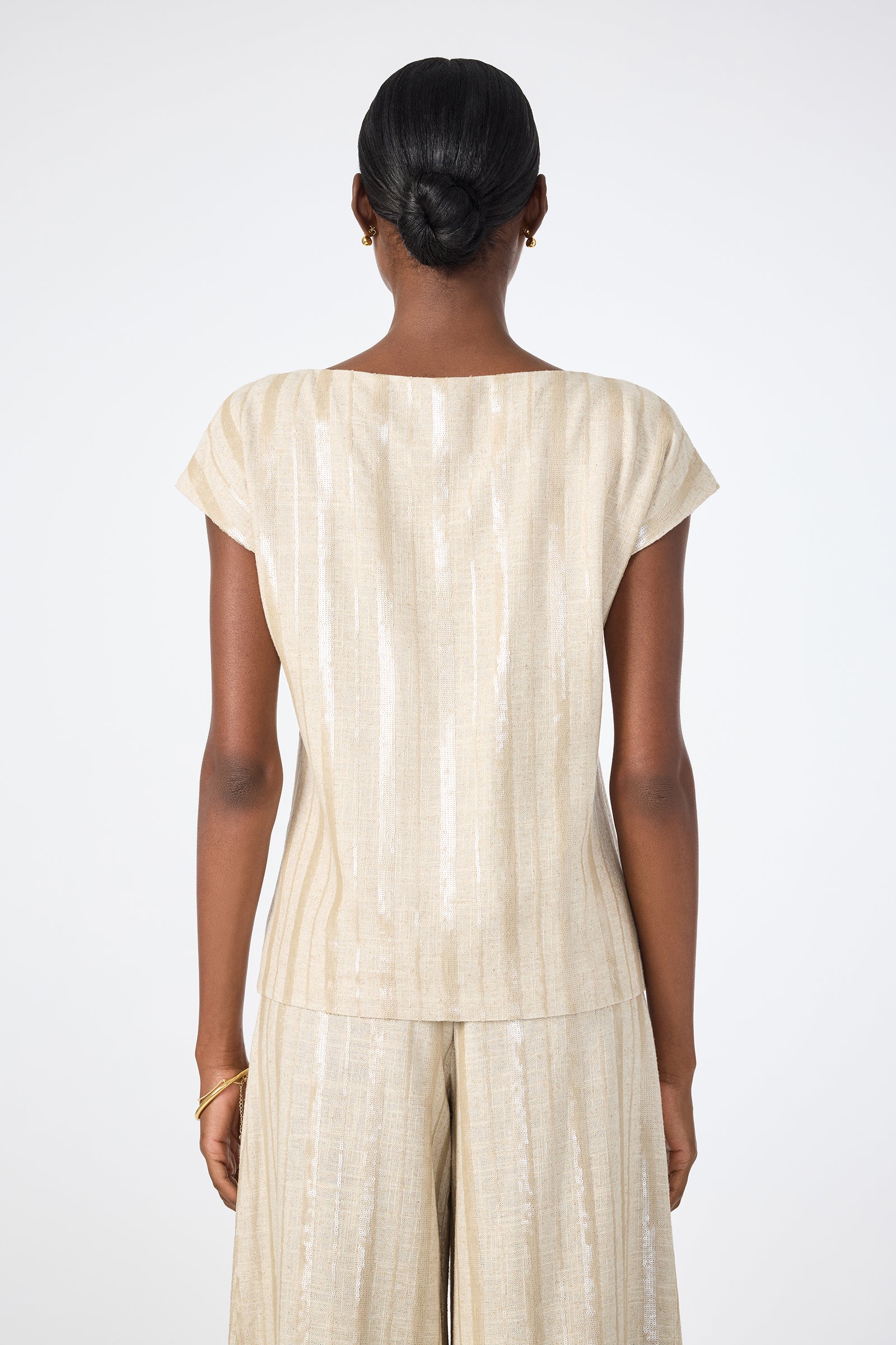 A woman with neatly styled hair in a bun is shown from behind, wearing the Breene Boatneck Top— a light-colored, short-sleeve piece paired with matching wide-leg trousers featuring a subtle striped pattern against a plain background.