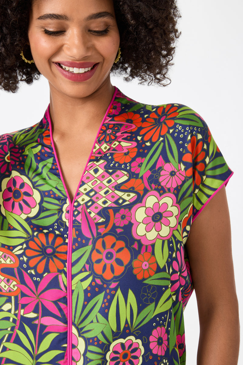 A smiling woman with curly hair models the Tarashi Short-Sleeve Mini Dress in Fumi Floral, featuring vibrant pink, green, orange, and navy patterns with a pink-trimmed neckline. Only her head and upper torso are shown against a white background.