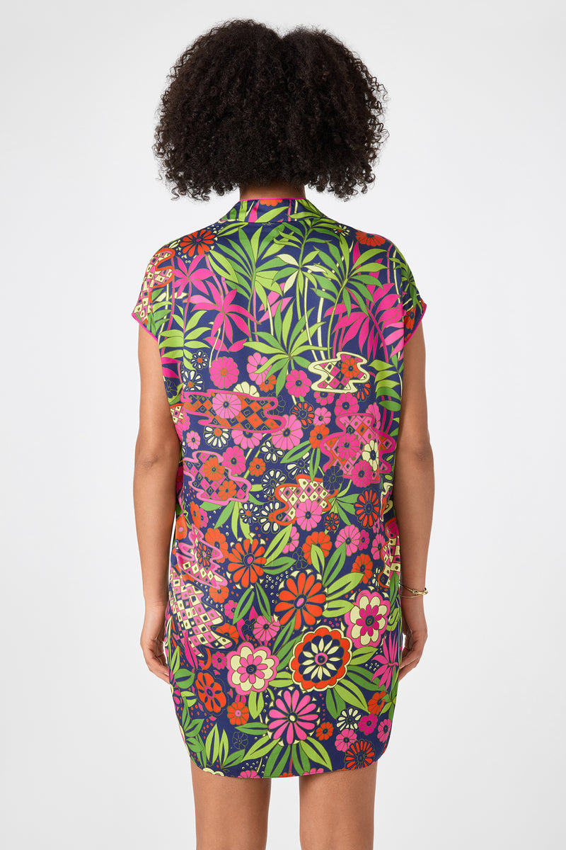 A person with curly hair is shown from the back, wearing the Tarashi Short-Sleeve Mini Dress - Fumi Floral, which features a bold and colorful floral pattern on a dark background.
