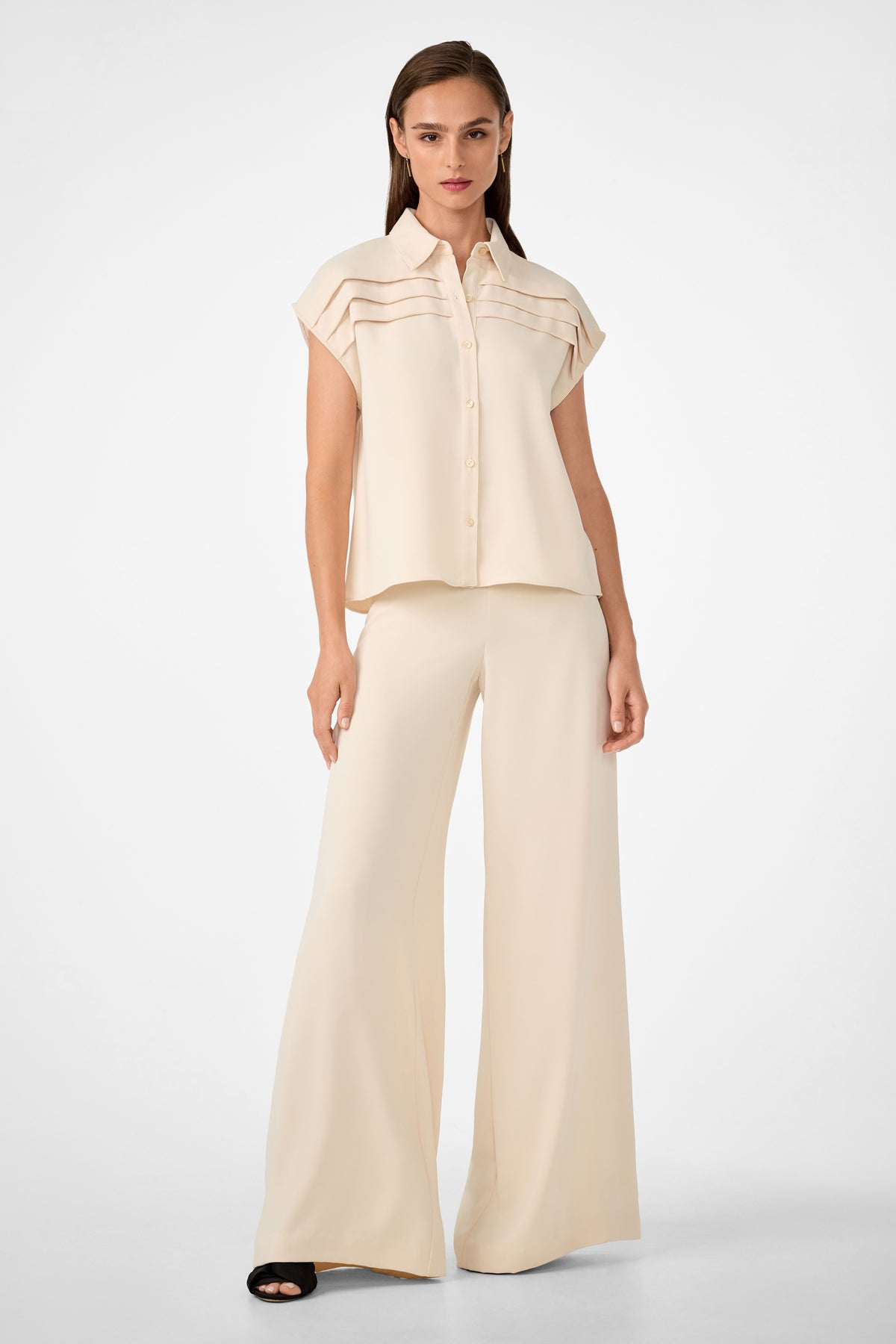 A woman stands against a plain white background wearing the Cheri Pleated Button-Front Top, a loose cream short-sleeve blouse with horizontal pleats, paired with wide-leg matching pants and black open-toe sandals.
