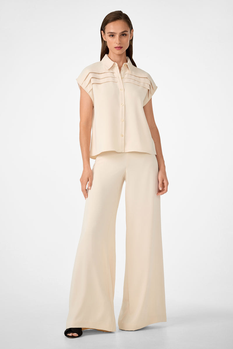 A woman stands against a plain white background wearing the Cheri Pleated Button-Front Top, a loose cream short-sleeve blouse with horizontal pleats, paired with wide-leg matching pants and black open-toe sandals.