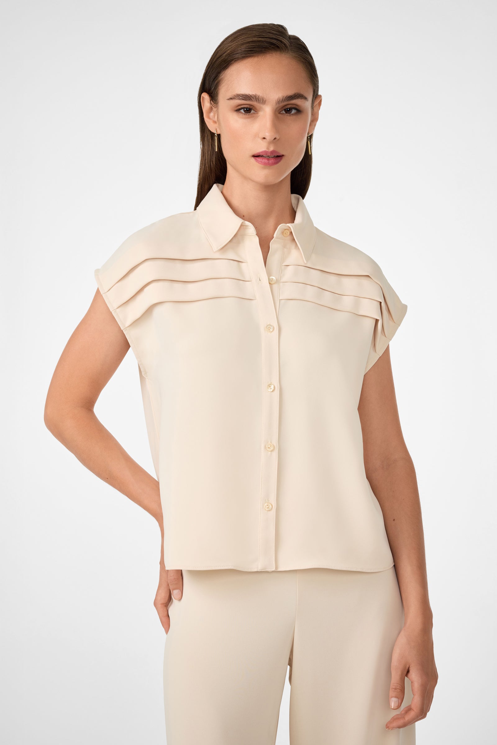 A woman wearing the Cheri Pleated Button-Front Top in Sancerre, featuring short sleeves and horizontal chest pleats, paired with matching beige pants, stands against a plain light background.