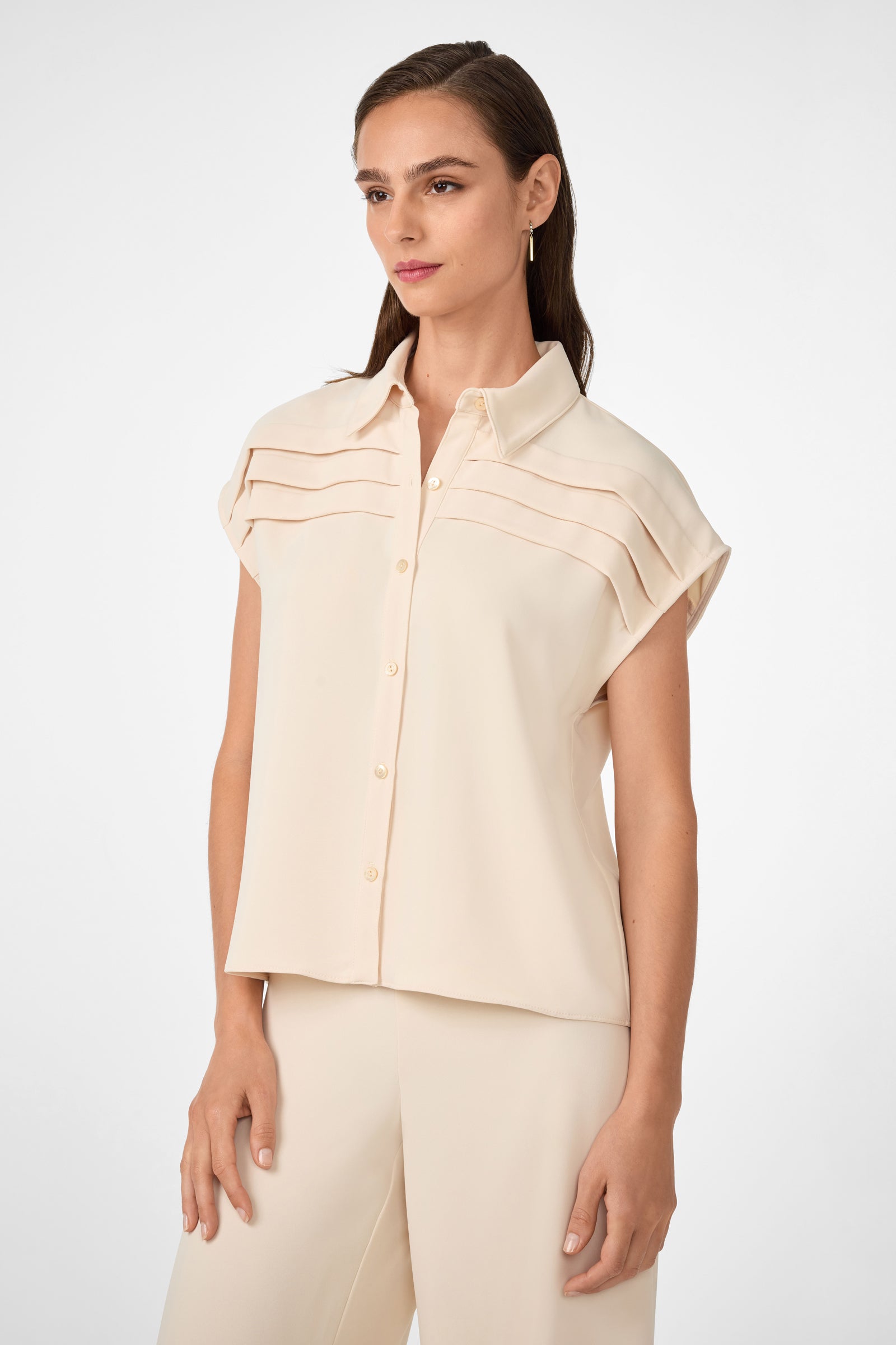 A woman wears the Cheri Pleated Button-Front Top—a short-sleeved, light beige blouse with pleated chest detail—and matching wide-leg pants, standing against a plain white background.