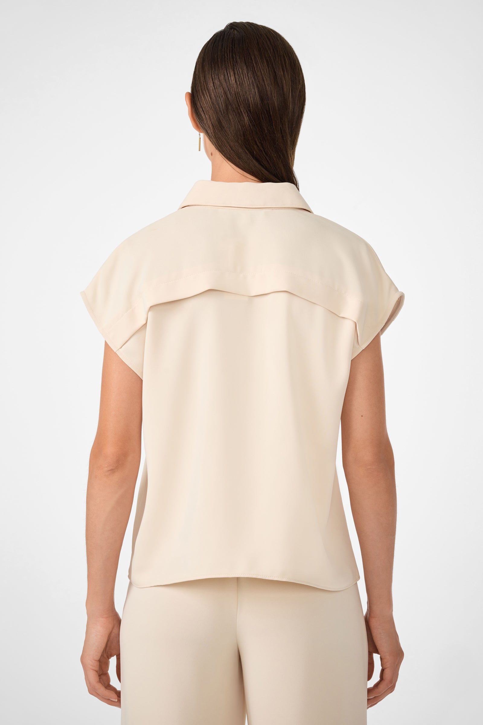 A woman, seen from the back, wears the Cheri Pleated Button-Front Top—a short-sleeve, light beige collared blouse—paired with matching beige pants. Her straight brown hair is tied back against a plain white background.