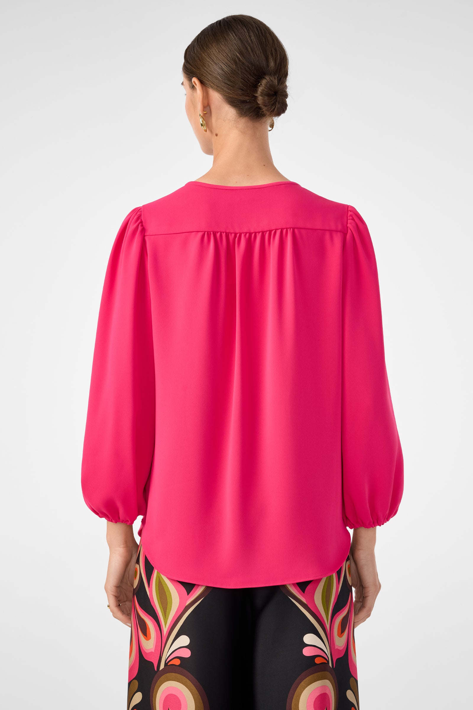 A woman with brown hair in a low bun is seen from behind wearing the Morabi Split-Neck Top in bright pink and colorful patterned pants. The background is plain and light-colored.