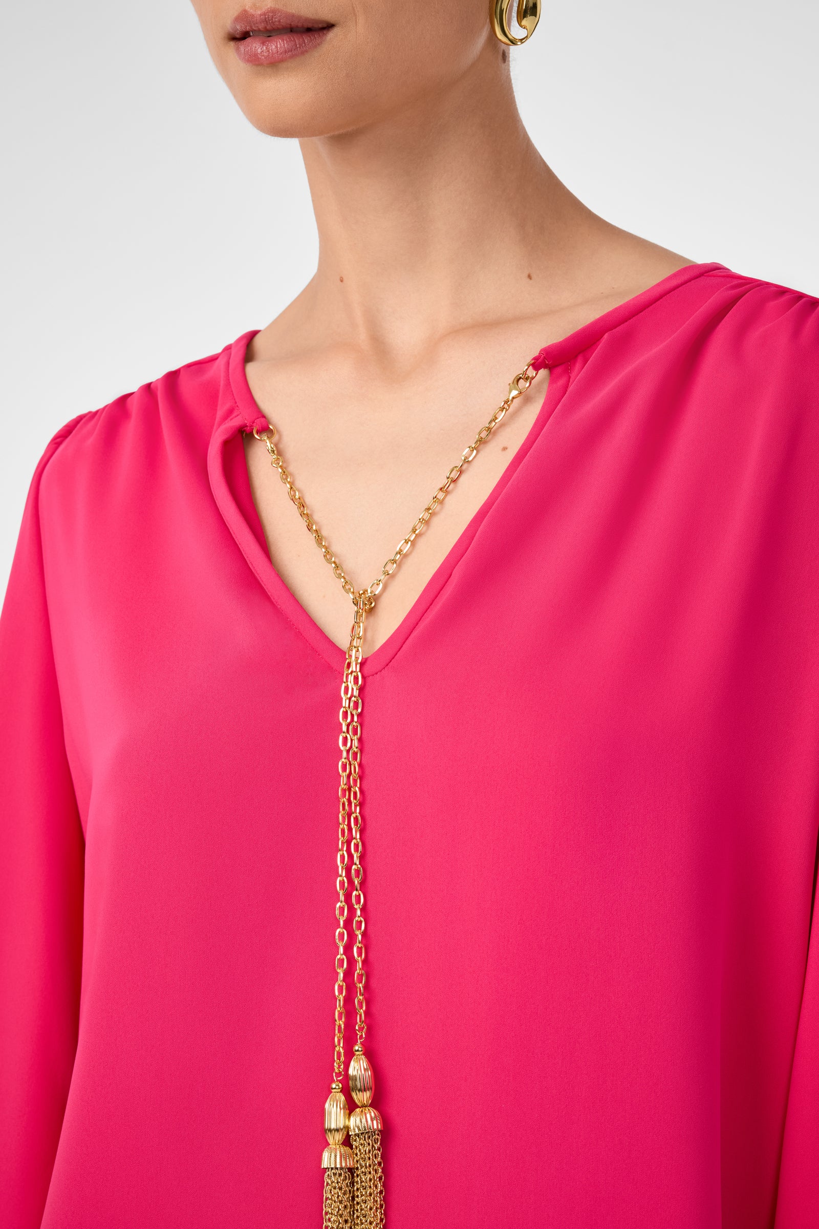 A woman wears the Morabi Split-Neck Top in bright pink, a gold chain necklace with decorative tassels, and a gold hoop earring. Her face is partially visible.