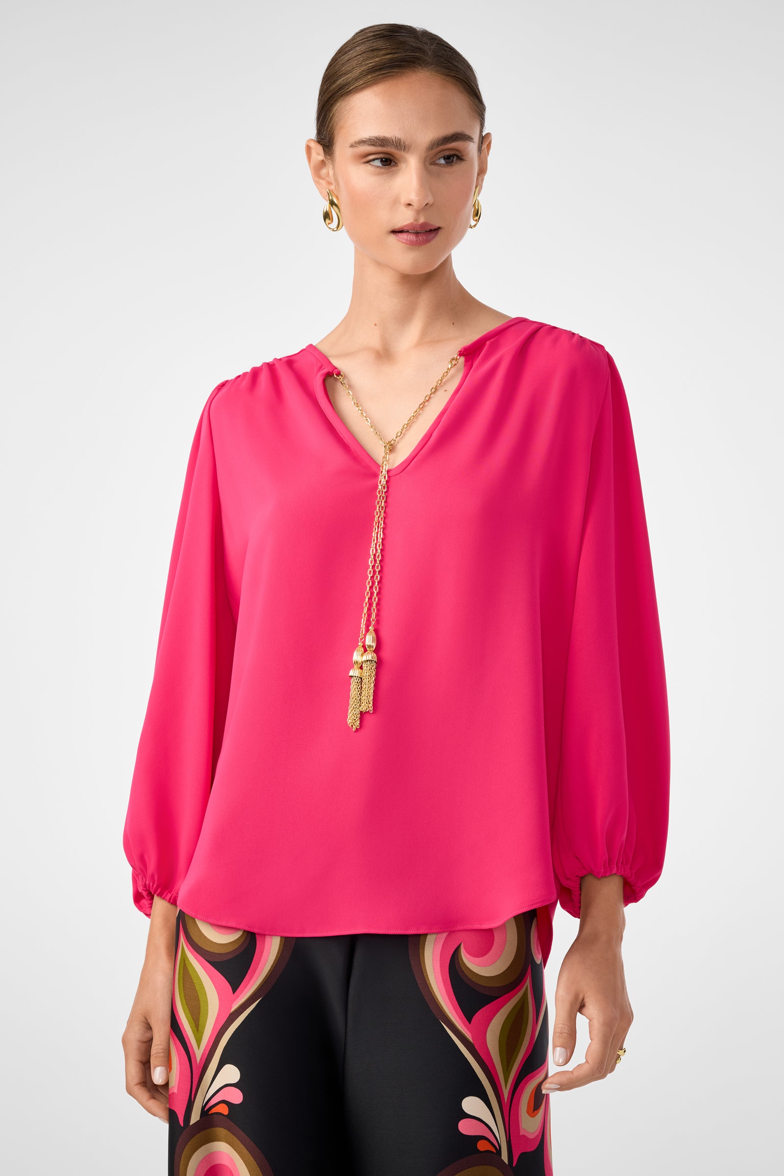 A woman wears the Morabi Split-Neck Top in Forrest Rose with a gold chain necklace and hoop earrings, pairing it with black pants that have colorful patterns at the waist. Her hair is pulled back.