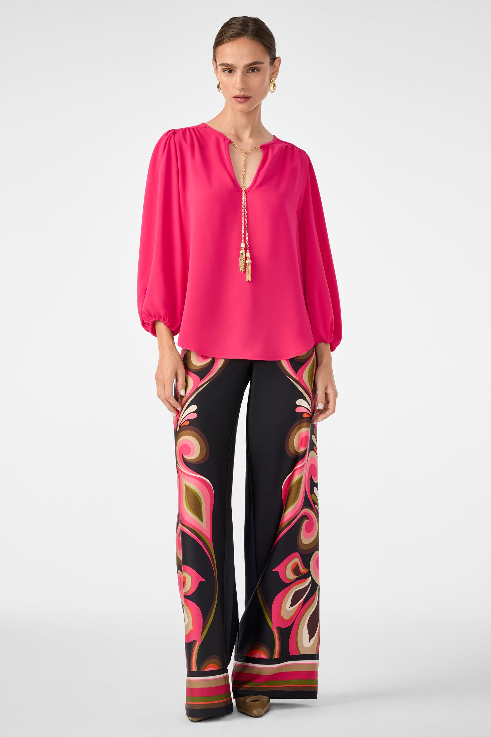 A woman wears the Morabi Split-Neck Top in bright pink, styled with a gold tassel necklace and high-waisted black flared pants covered in bold, colorful abstract patterns.