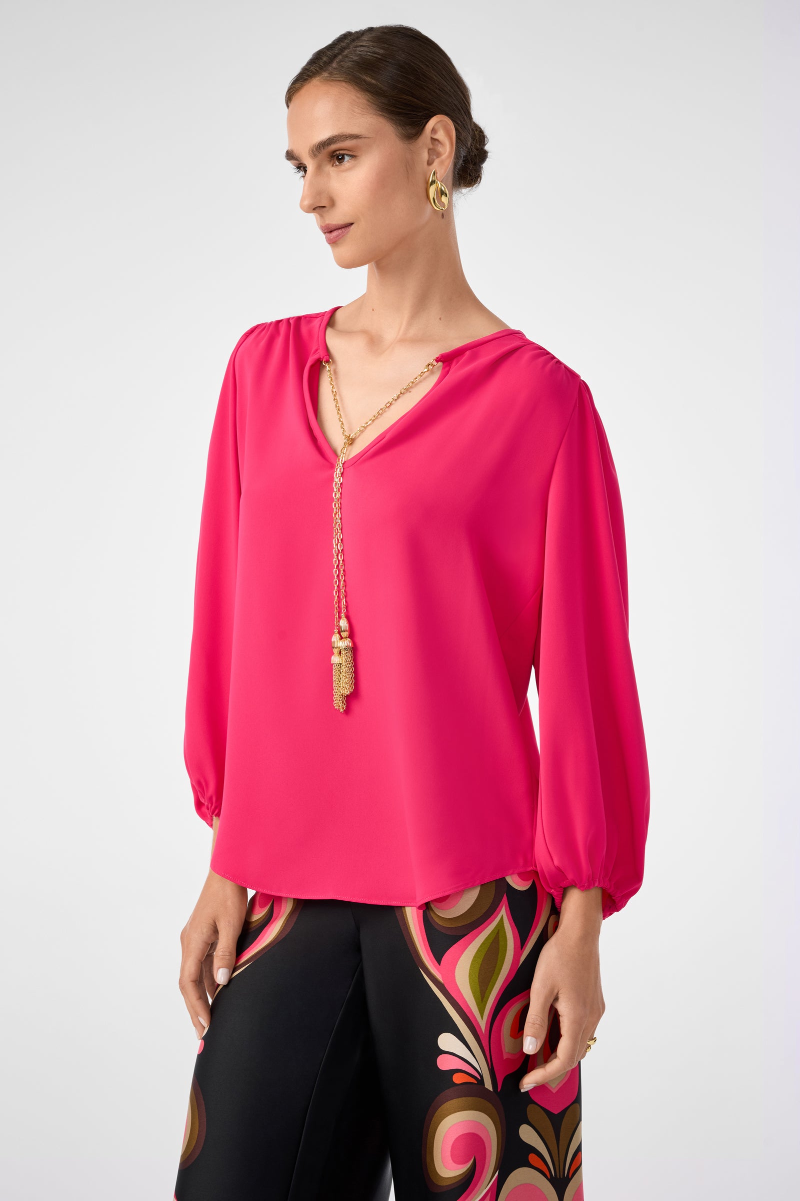 A woman wears the Morabi Split-Neck Top in bright pink with a gold chain necklace and black pants featuring a bold, colorful swirl pattern, standing against a plain light background.