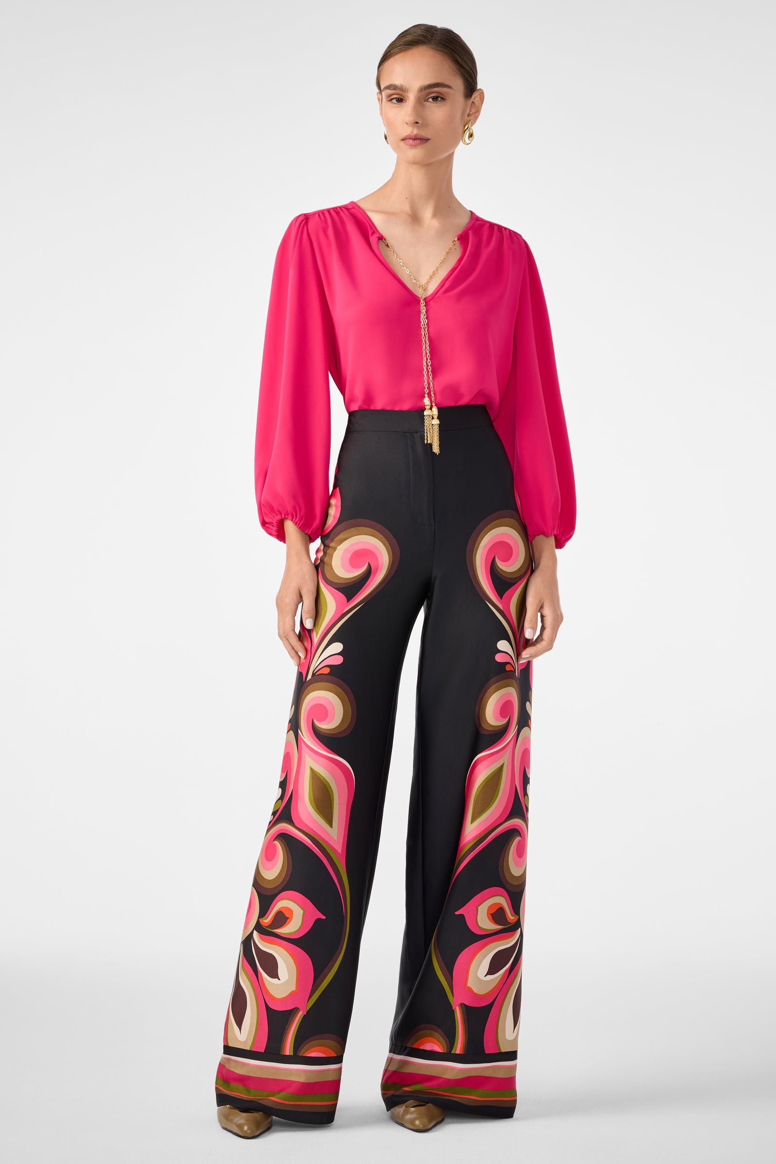 A woman wears the Morabi Split-Neck Top in bright pink with balloon sleeves, paired with high-waisted black pants featuring pink, orange, and cream abstract patterns. She accessorizes with gold earrings and brown pointed shoes.