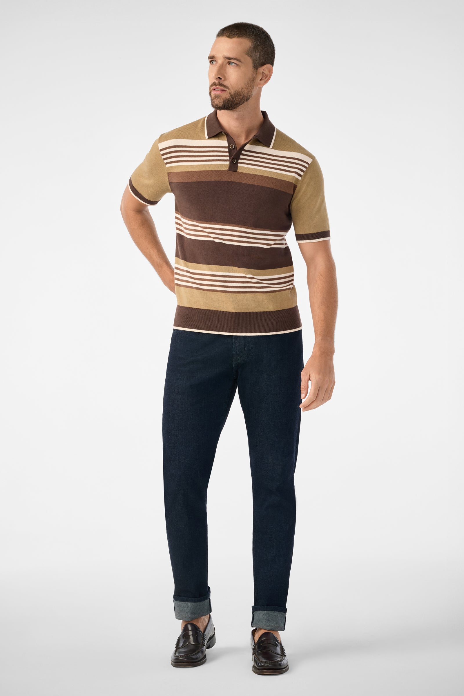 A man with short hair and a beard stands against a plain background, wearing the Edward Short-Sleeve Polo in brown and tan stripes, dark cuffed jeans, and brown loafers. He looks slightly to his right.