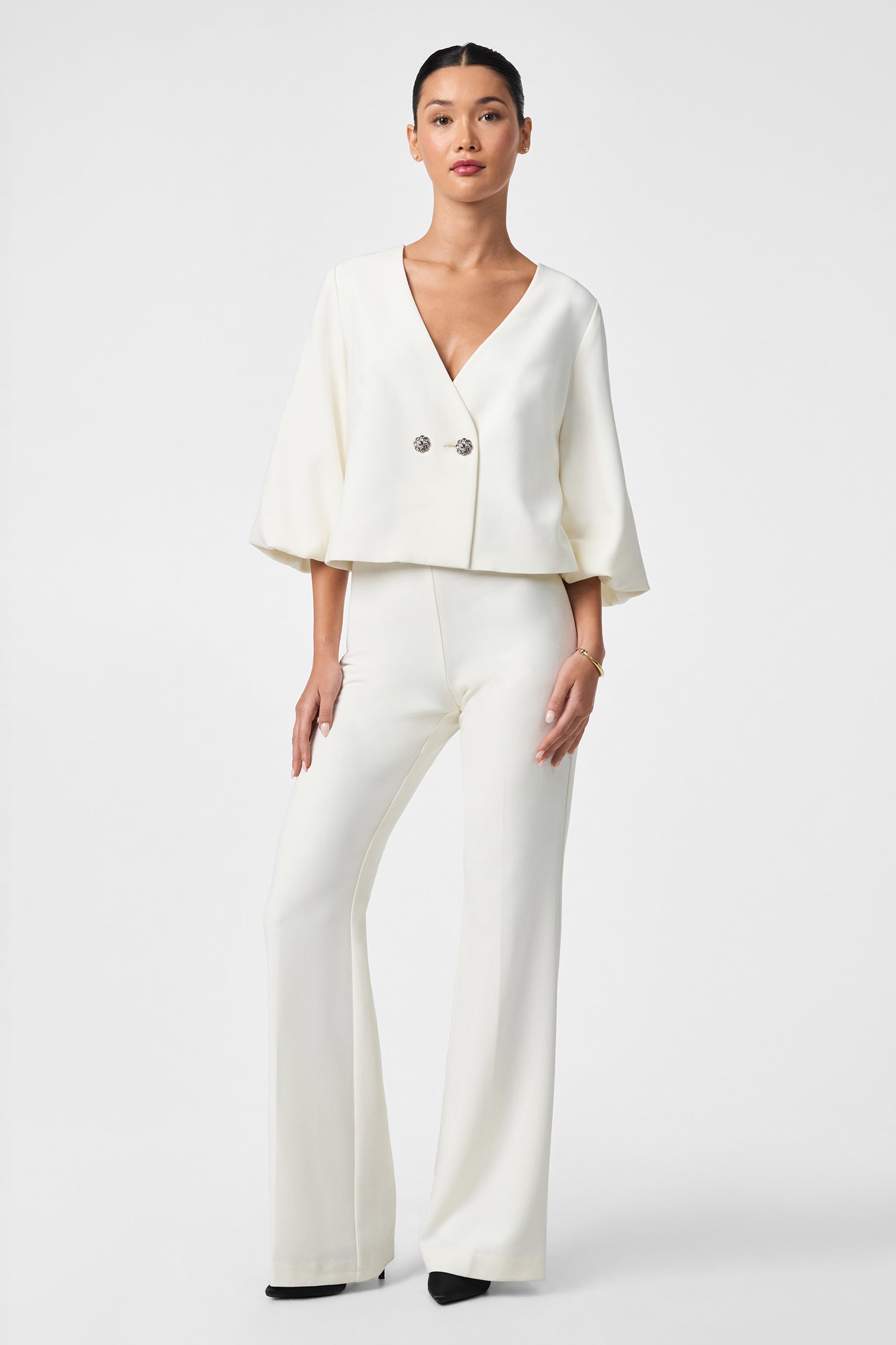 A woman poses against a plain white background wearing the Basal Cropped Jacket with flared sleeves and two buttons, styled with matching wide-leg pants and black pointed-toe shoes.