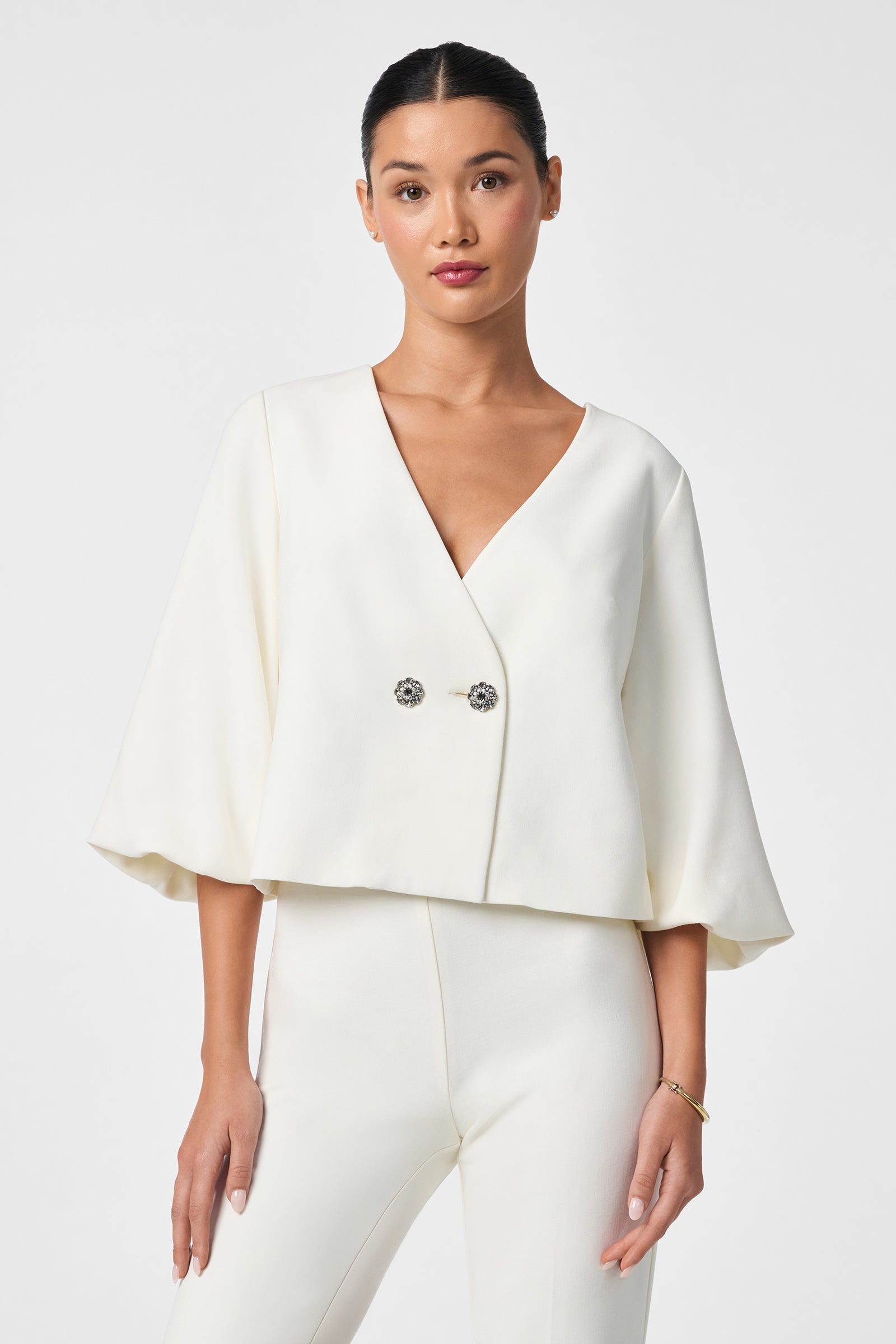 A woman with her hair tied back and a neutral expression models the Basal Cropped Jacket—a white wide-sleeve blazer with two decorative buttons—paired with matching white pants against a plain light background.