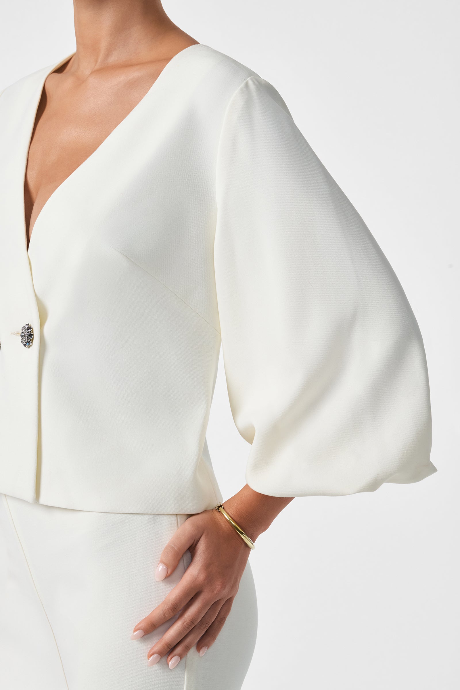 A woman wears the Basal Cropped Jacket, an elegant white suit with wide, billowy sleeves, a deep V-neckline, and decorative buttons. She accessorizes with a simple gold bracelet against a plain white background.