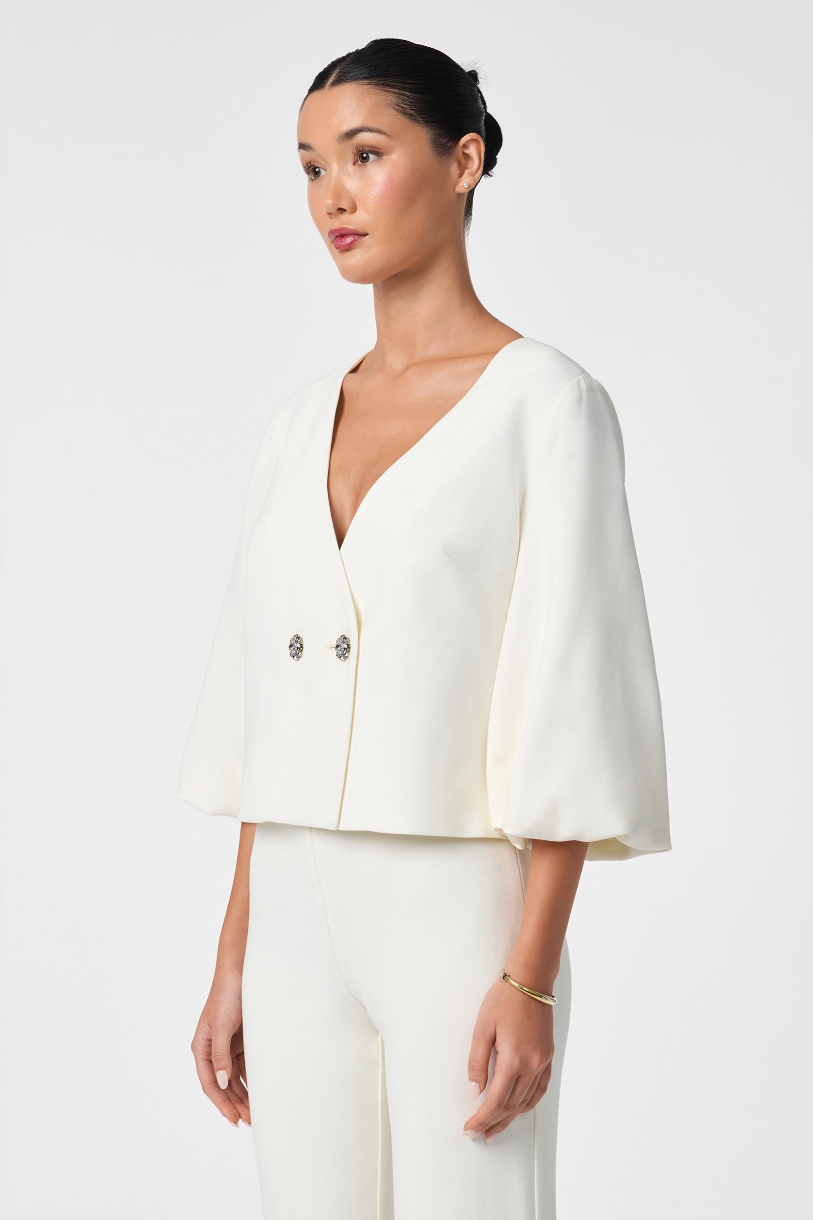 A woman with dark hair in a bun models the Basal Cropped Jacket, a stylish white suit with wide three-quarter sleeves and two decorative buttons, paired with matching white pants against a plain light background.
