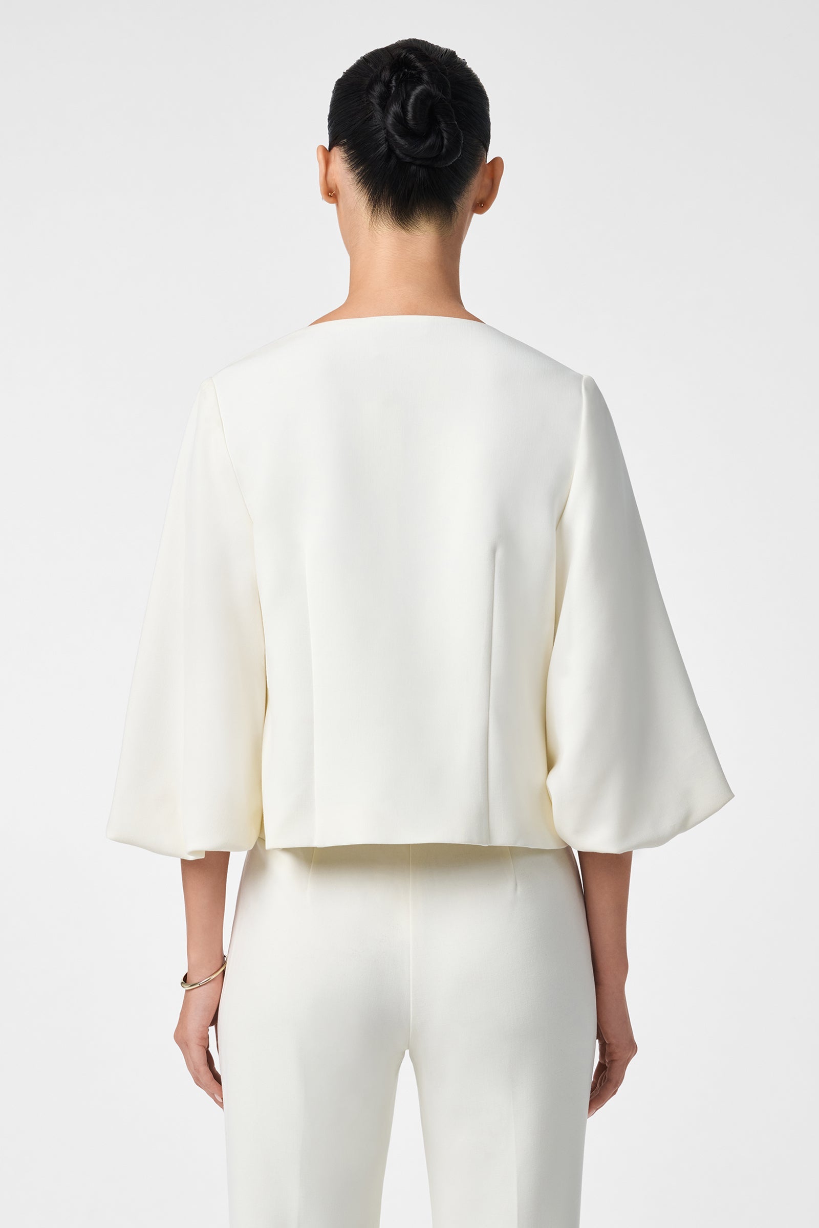 A person with dark hair in a low bun is shown from the back, wearing the off-white Basal Cropped Jacket with three-quarter sleeves and matching pants, set against a plain white background.