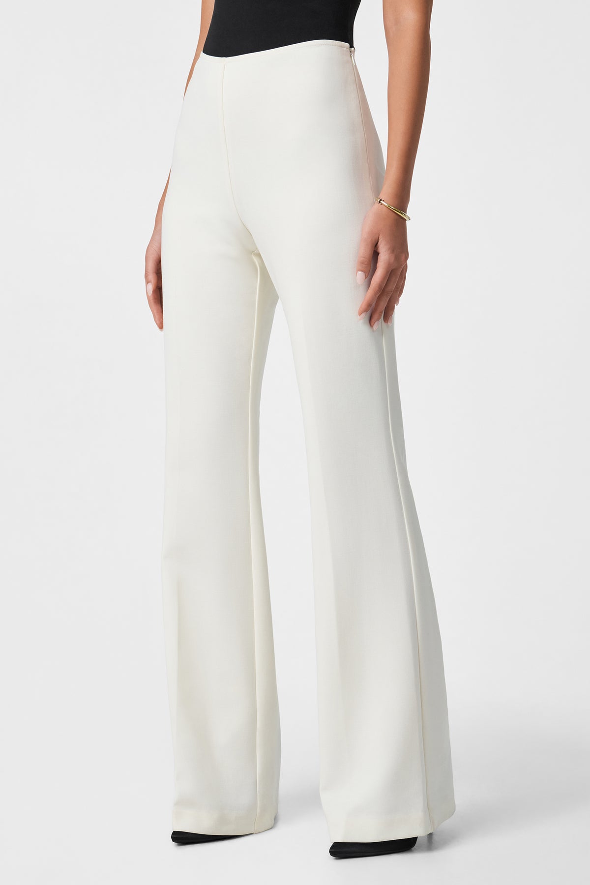 A person wearing Chimayo 2 Pant—high-waisted, wide-leg white pants—paired with a black sleeveless top, gold bracelet, and black pointed-toe shoes, stands against a plain white background.