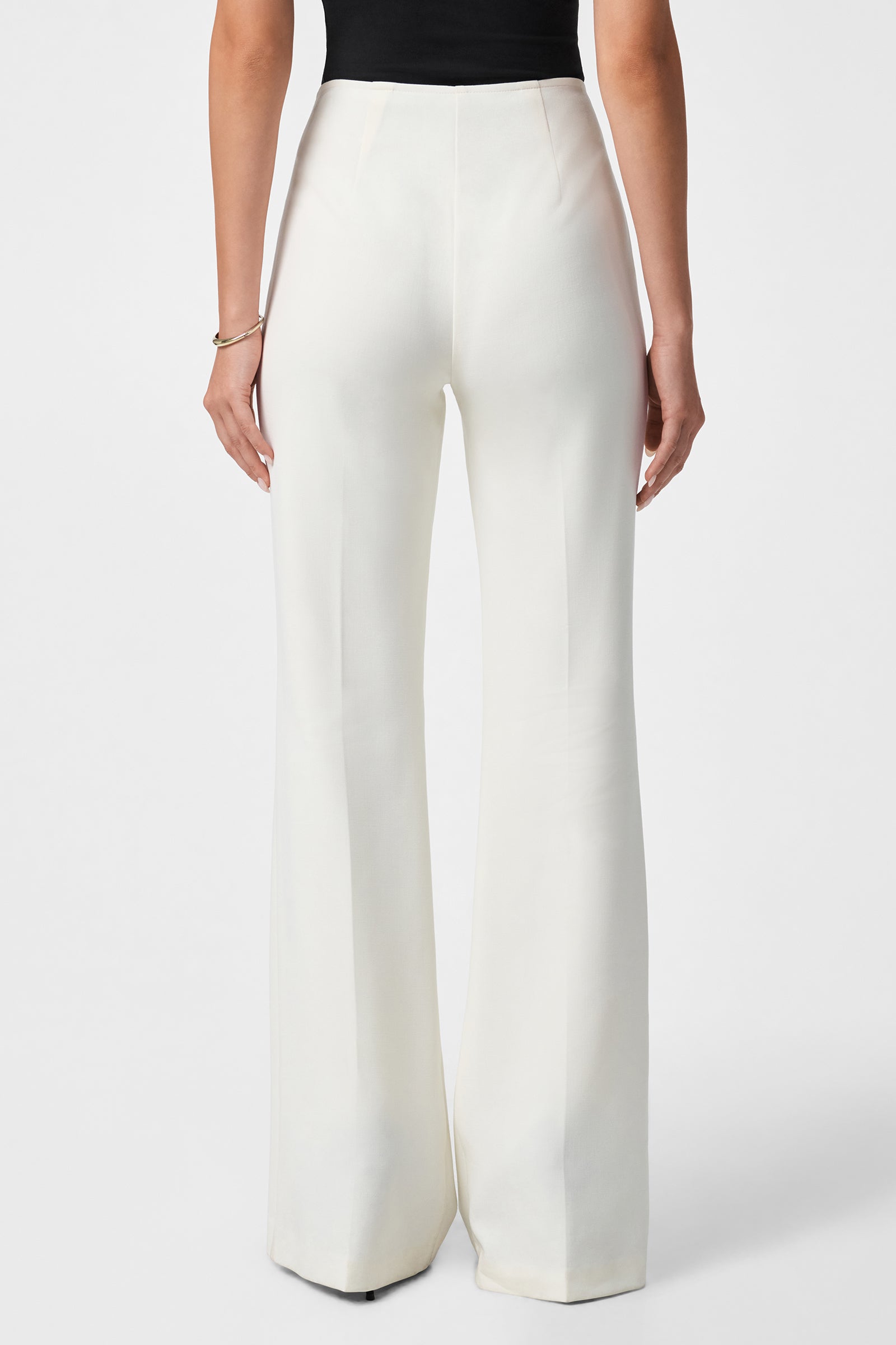 A person stands facing away, wearing the Chimayo 2 Pant—high-waisted, wide-leg white trousers with a smooth finish and flowing legs—paired with a black sleeveless top against a plain white background.