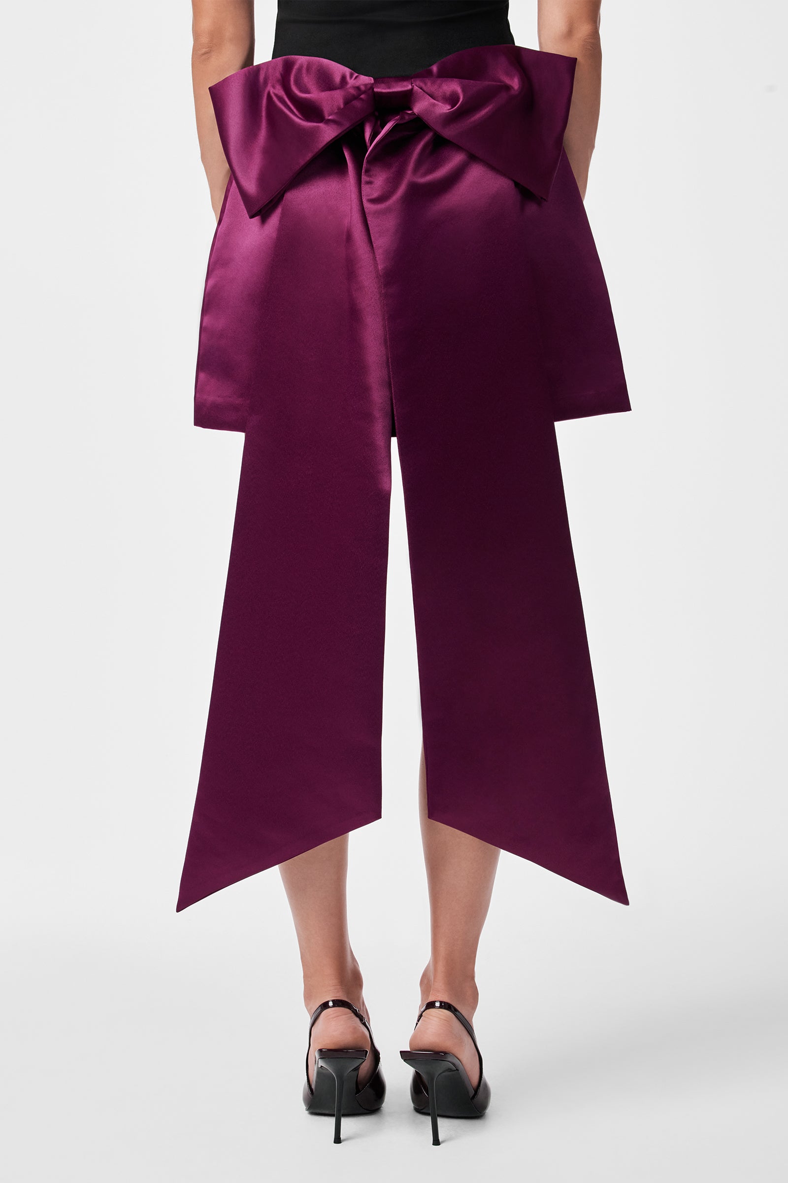 A person wears the Vinna Skirt—a large, deep purple satin bow skirt with long, pointed tails at the back—paired with a black top and black heels, elegantly draping to mid-calf.