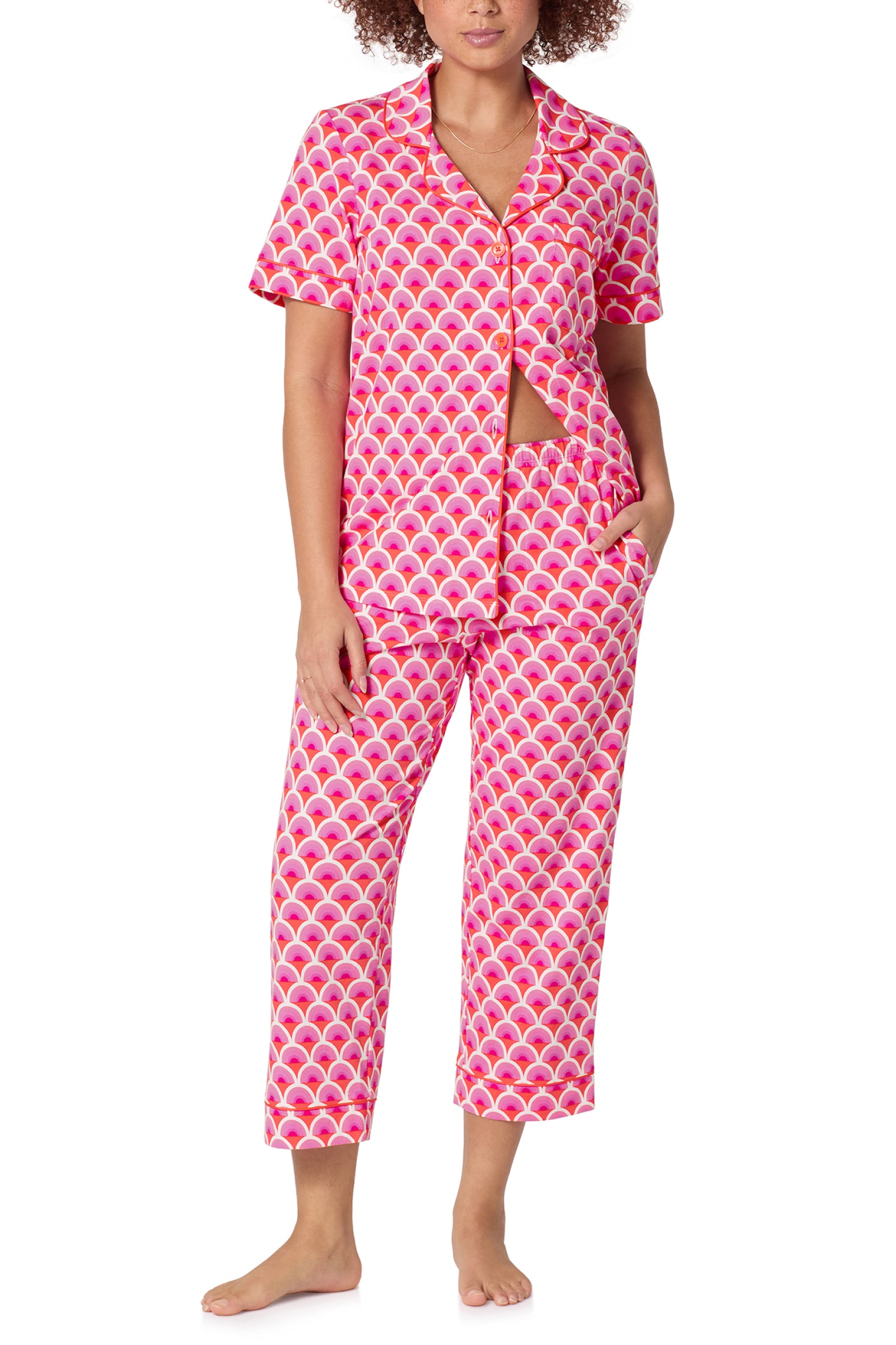 A person wearing the Crop Classic PJ Set - Scallop, featuring a pink and white scallop pattern with a short-sleeve button-up top and cropped pants, stands barefoot against a white background.