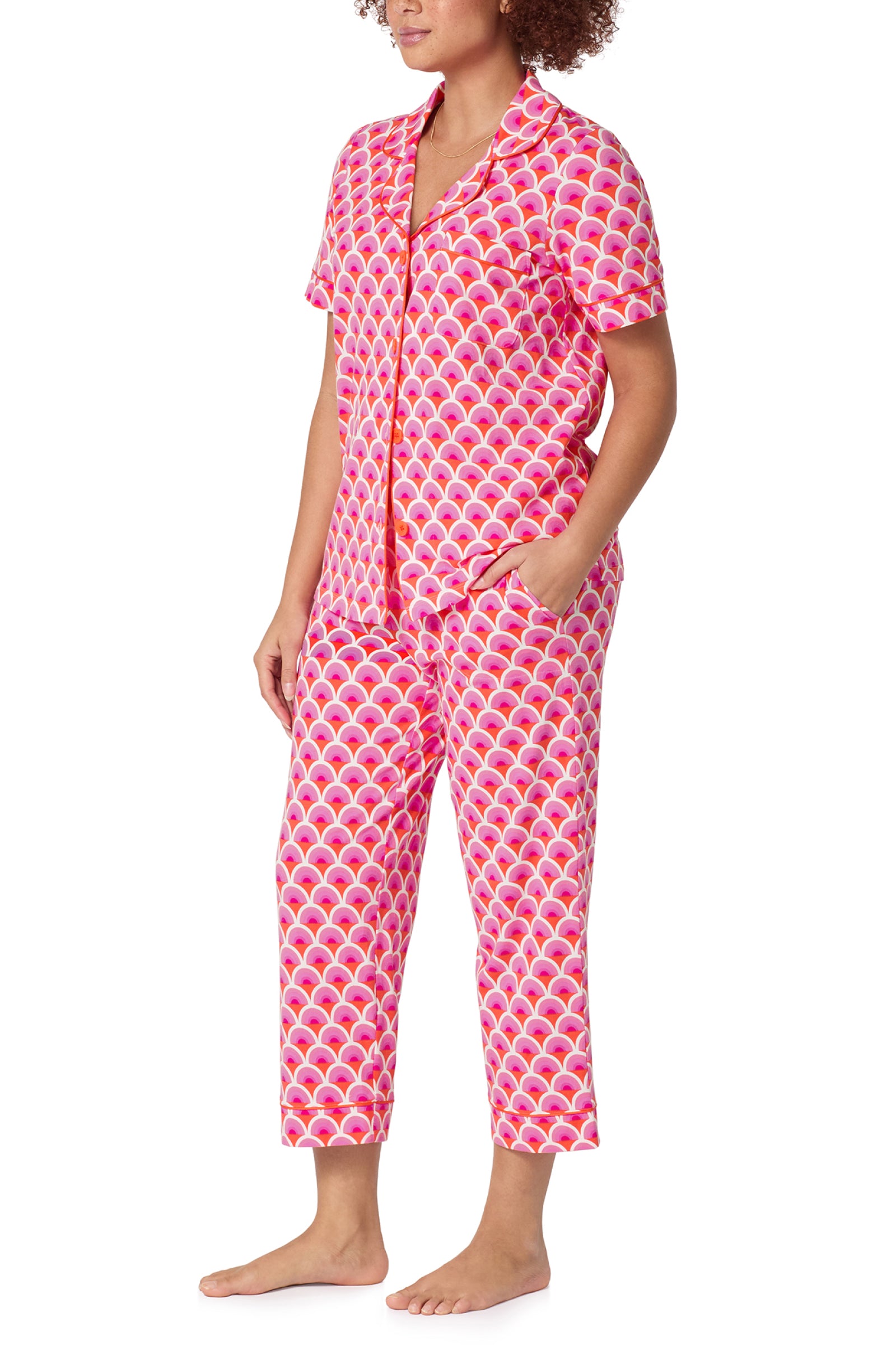 Someone stands barefoot in the Crop Classic PJ Set - Scallop, featuring a bold pink and orange scallop pattern with a short-sleeve button-up top and cropped pants with pockets.