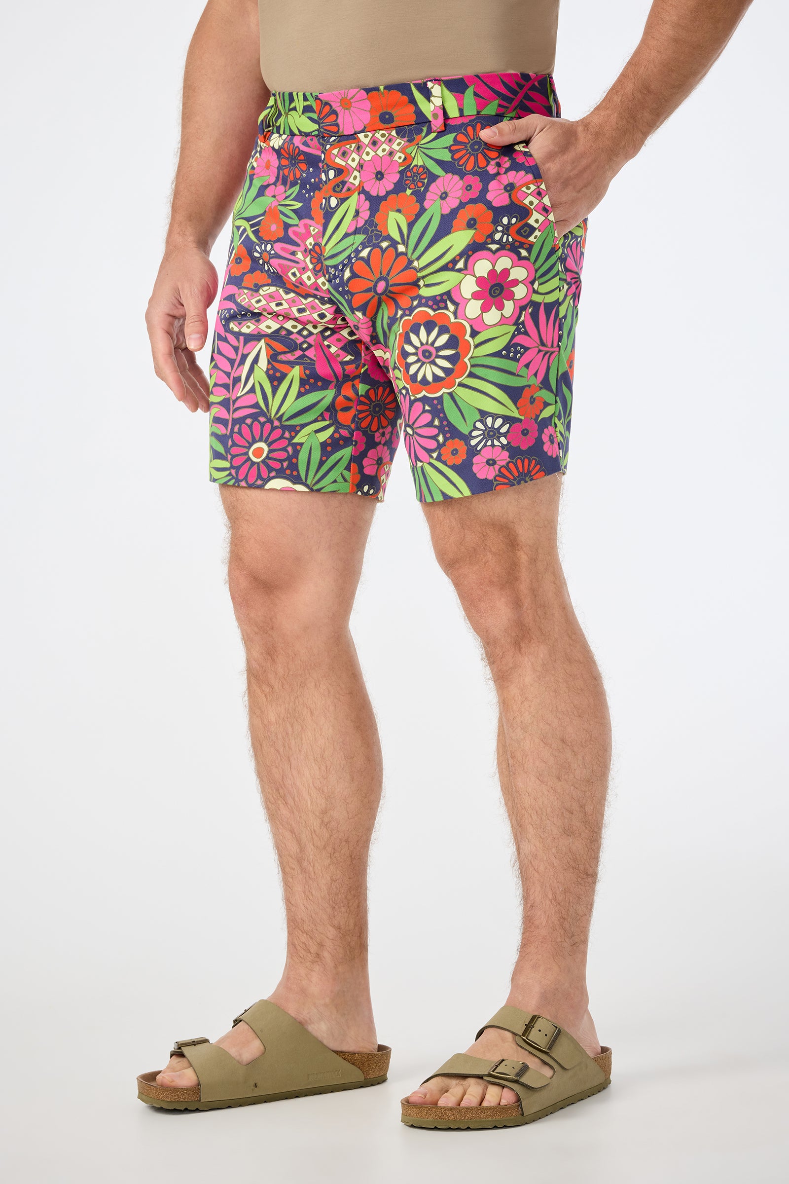 A man stands against a plain white background, wearing the Lawrence Short - Fumi Floral with a beige shirt and tan sandals, one hand in his pocket.