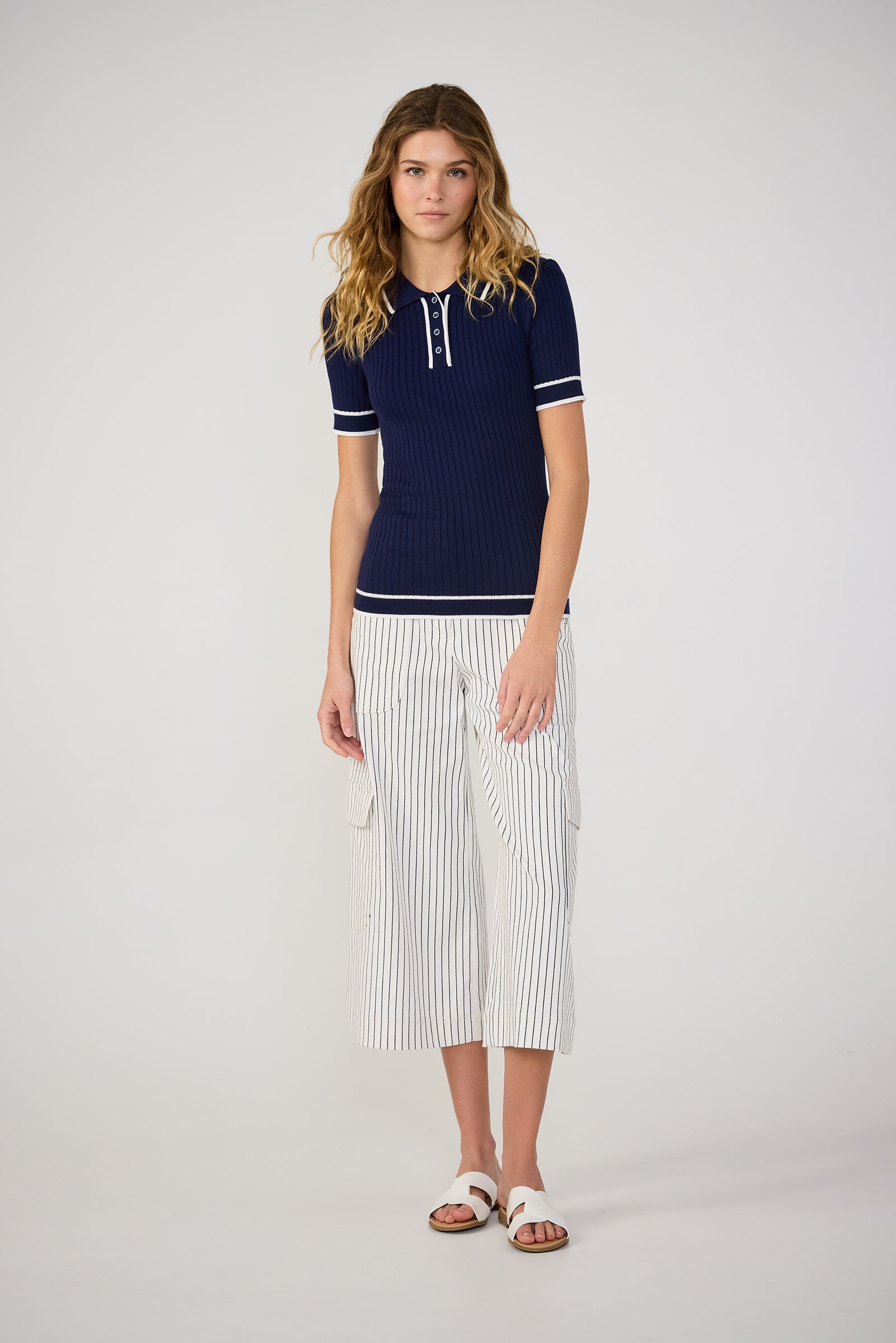 A woman stands against a plain background wearing the Stance Contrast Trim Short-Sleeve Polo in navy with white trim, paired with white wide-leg striped cropped pants and white sandals.