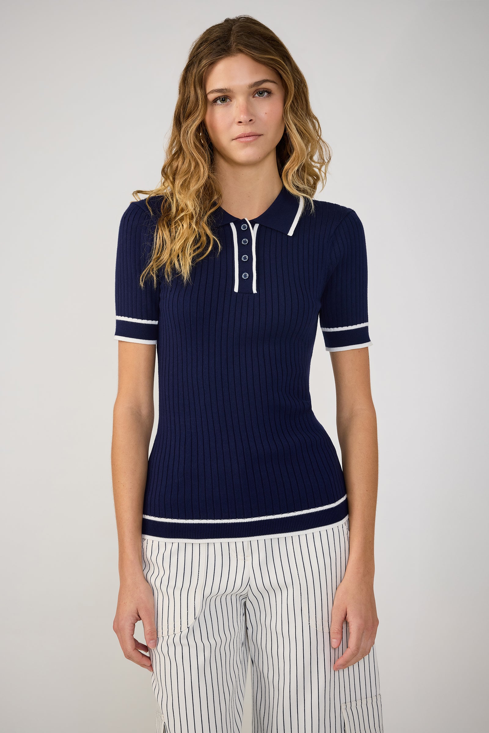 A woman with wavy hair wears the Stance Contrast Trim Short-Sleeve Polo in Ink/Whitewash, paired with white striped pants, standing against a plain light background.