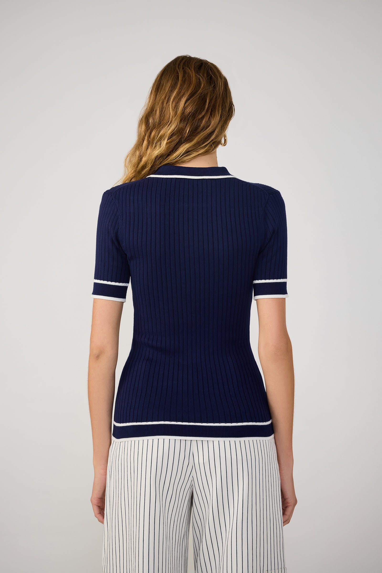 A woman with wavy, light brown hair is seen from the back, wearing the Stance Contrast Trim Short-Sleeve Polo in navy blue with white trim and white pants with thin black vertical stripes. The background is plain and light.