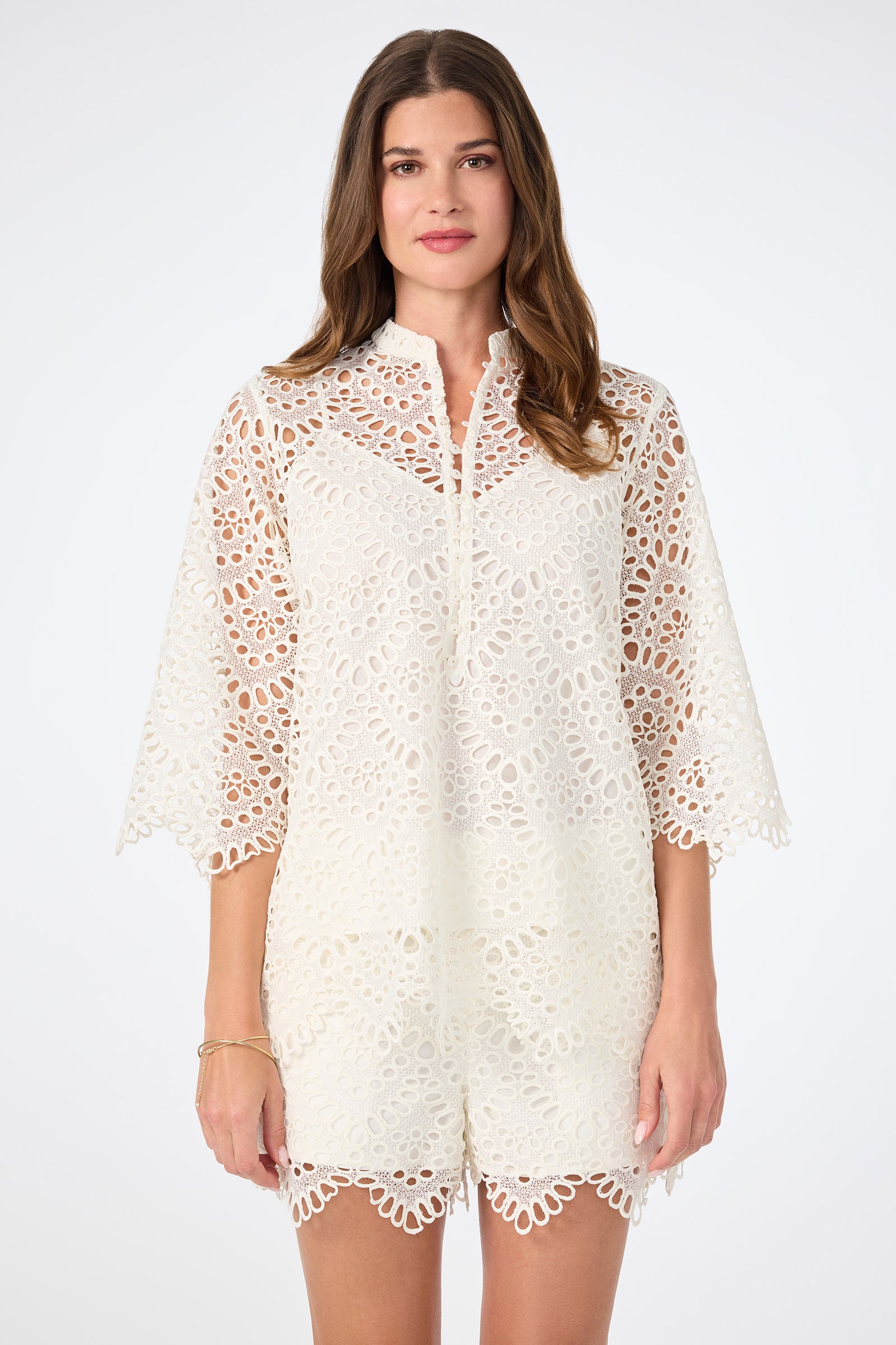 A woman with long brown hair wears the Martigues Lace Top, featuring a white lace pattern, scalloped edges, and wide sleeves, against a plain white background.