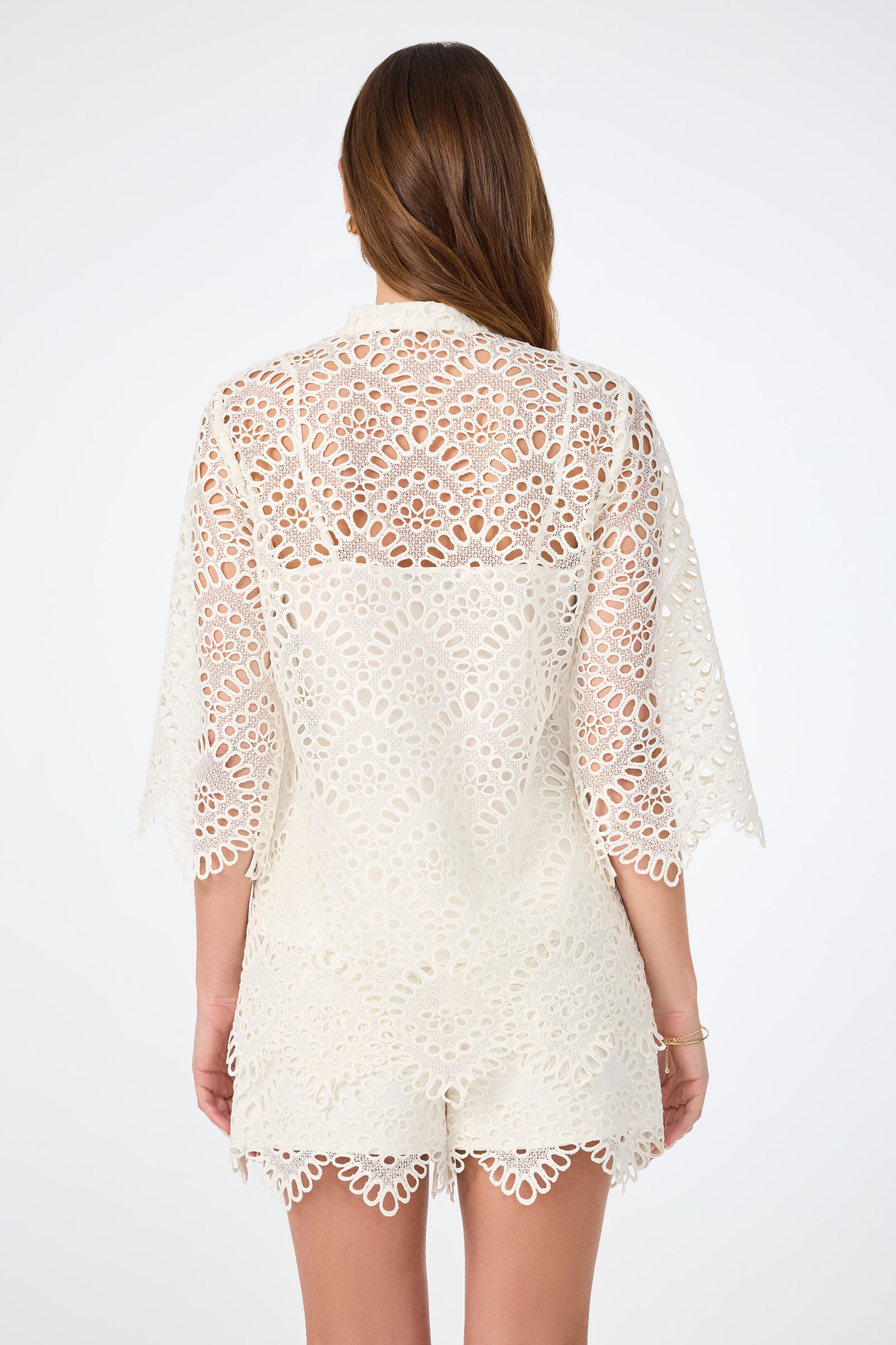 A woman with long brown hair faces away, wearing the Martigues Lace Top and matching shorts featuring scallop edges and intricate cutout patterns. The background is plain white.