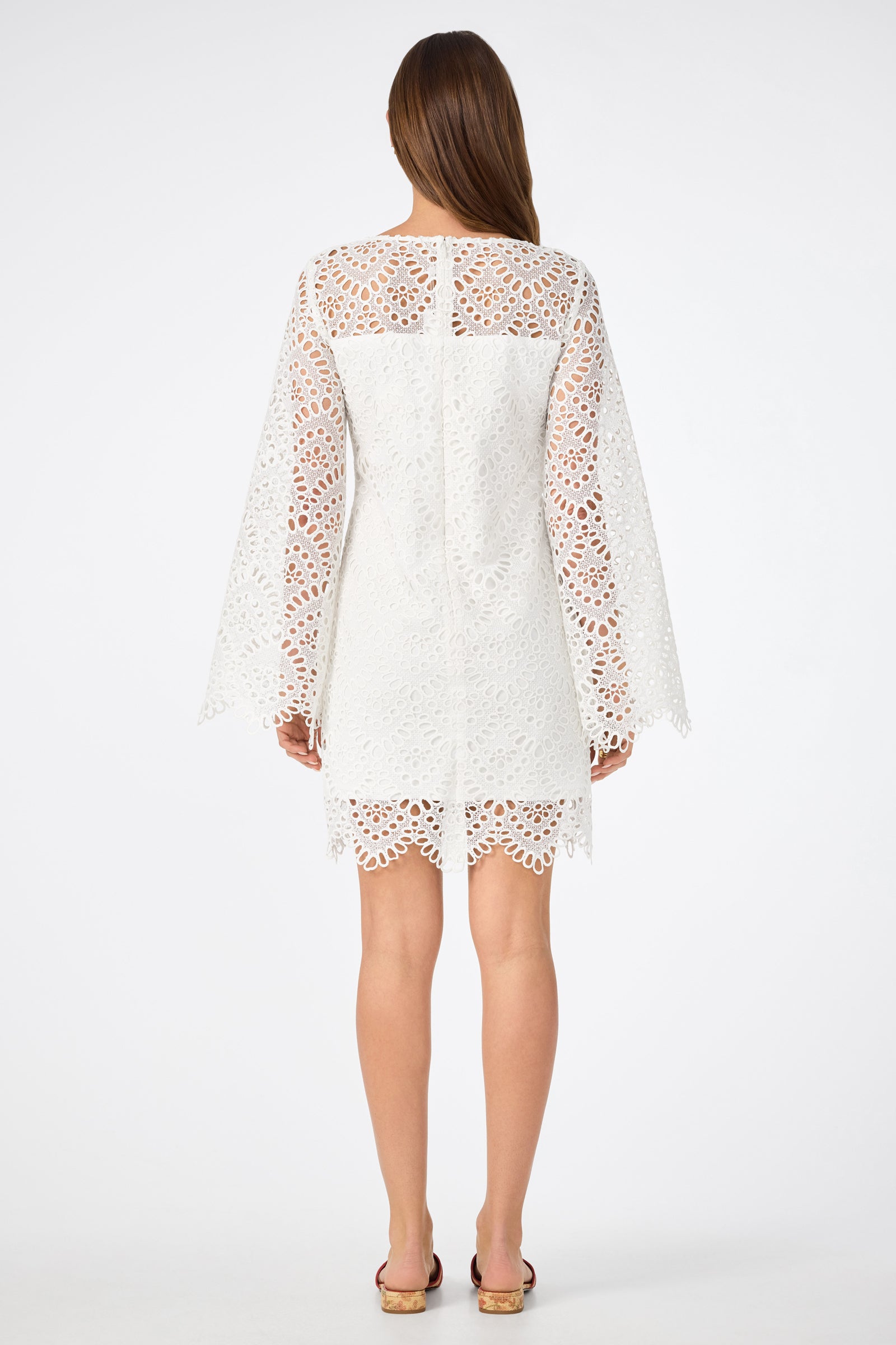 A woman faces away, wearing the Arles Dress—a white, knee-length lace dress with wide long sleeves and intricate cut-out patterns. She has straight brown hair and open-toe sandals, standing against a plain white background.