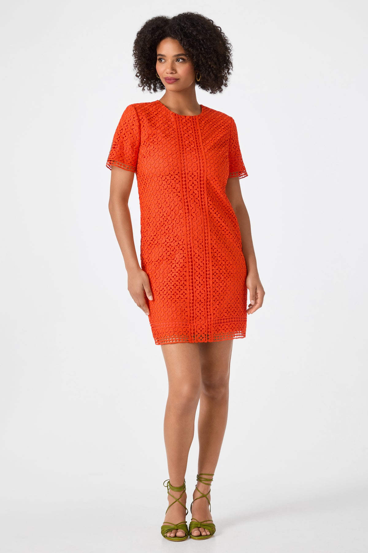 A woman with curly hair wears the Kimasu Short-Sleeve Lace Mini Dress in Ryuji Red, paired with green strappy sandals, standing against a plain white background.