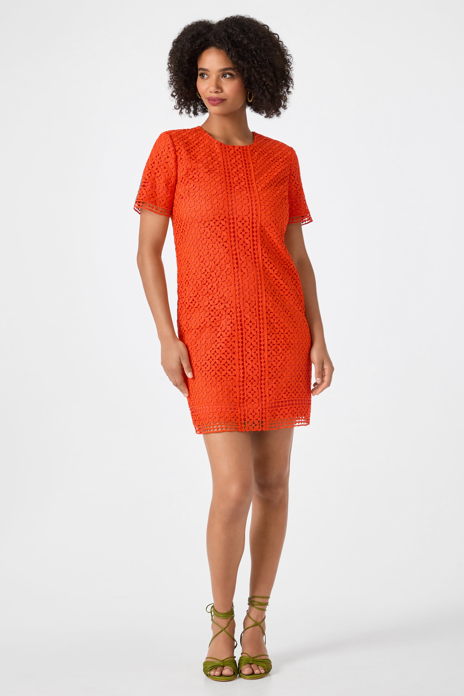 A woman with curly hair wears the Kimasu Short-Sleeve Lace Mini Dress in Ryuji Red, paired with green strappy sandals, standing against a plain white background.