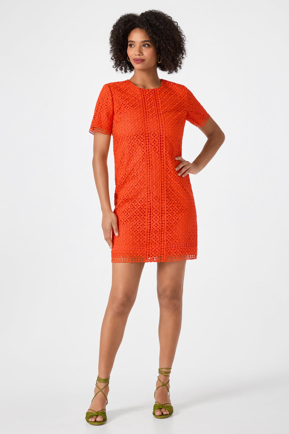 A woman stands confidently in the Kimasu Short-Sleeve Lace Mini Dress, a bright orange shift silhouette. She pairs it with green strappy heels and wears her natural curly hair. The background is plain white.
