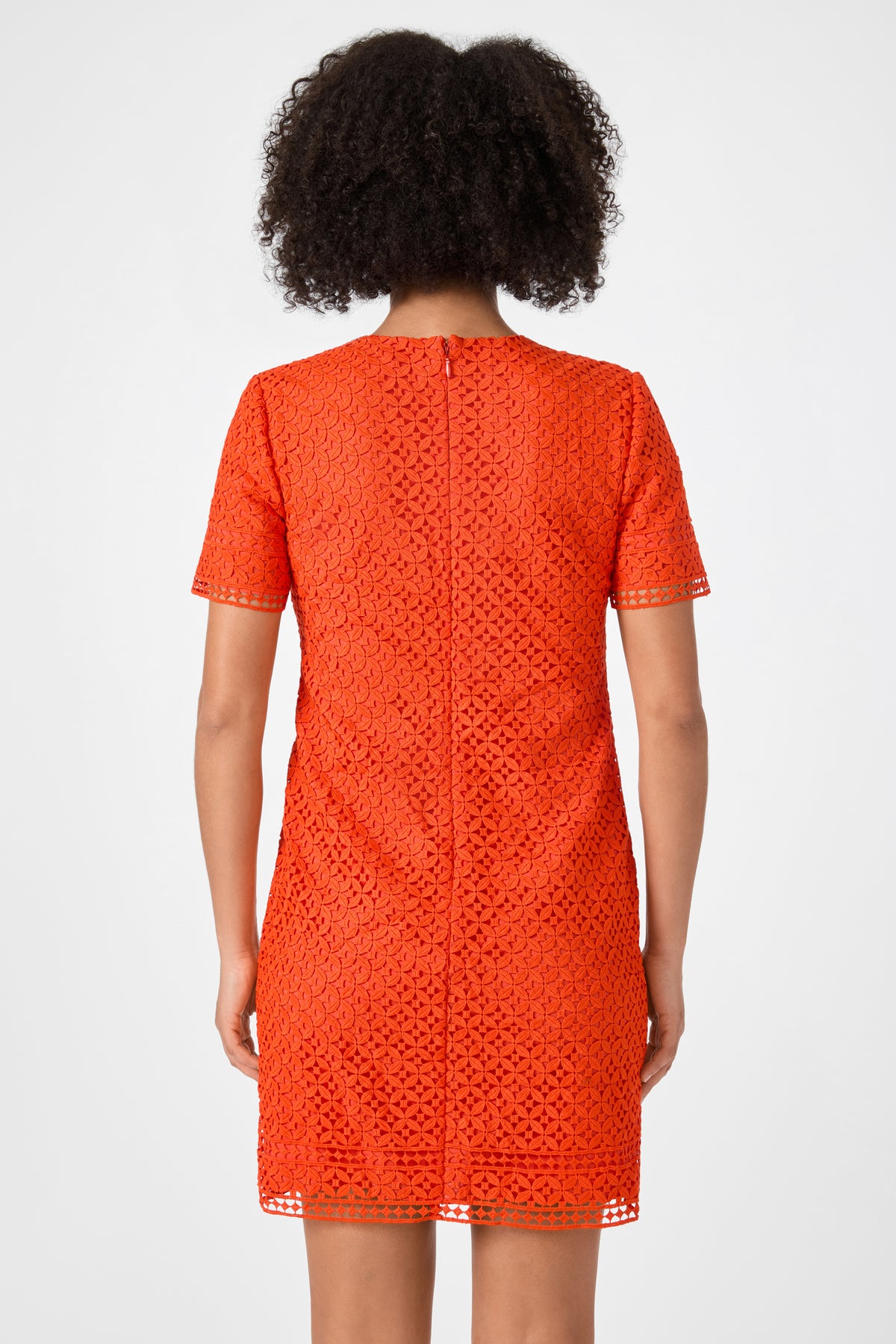 A person with curly dark hair is shown from the back, wearing the Kimasu Short-Sleeve Lace Mini Dress in bright orange. The knee-length dress features a geometric lace pattern, short sleeves, and a visible central seam.
