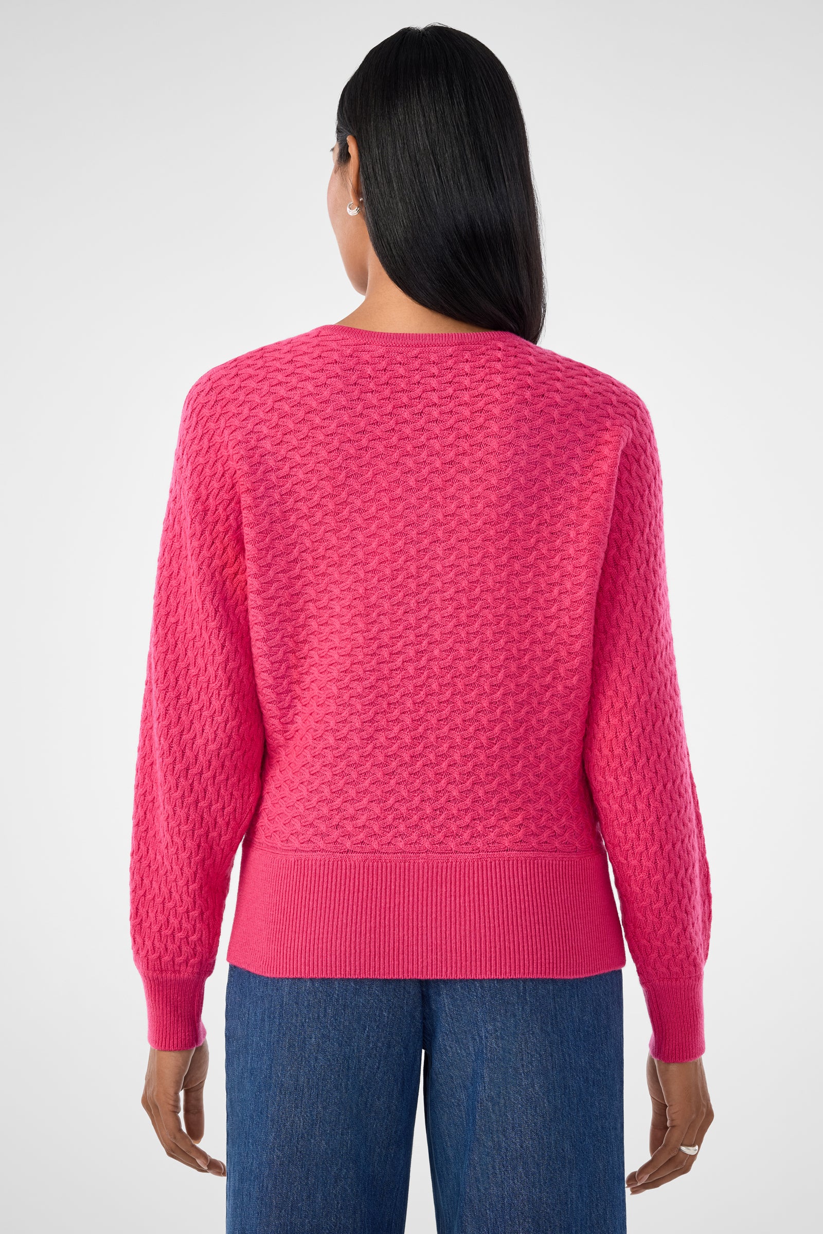A person with long, straight black hair is seen from the back, wearing the Lima V-Neck Pullover in bright pink and blue jeans against a plain light background.