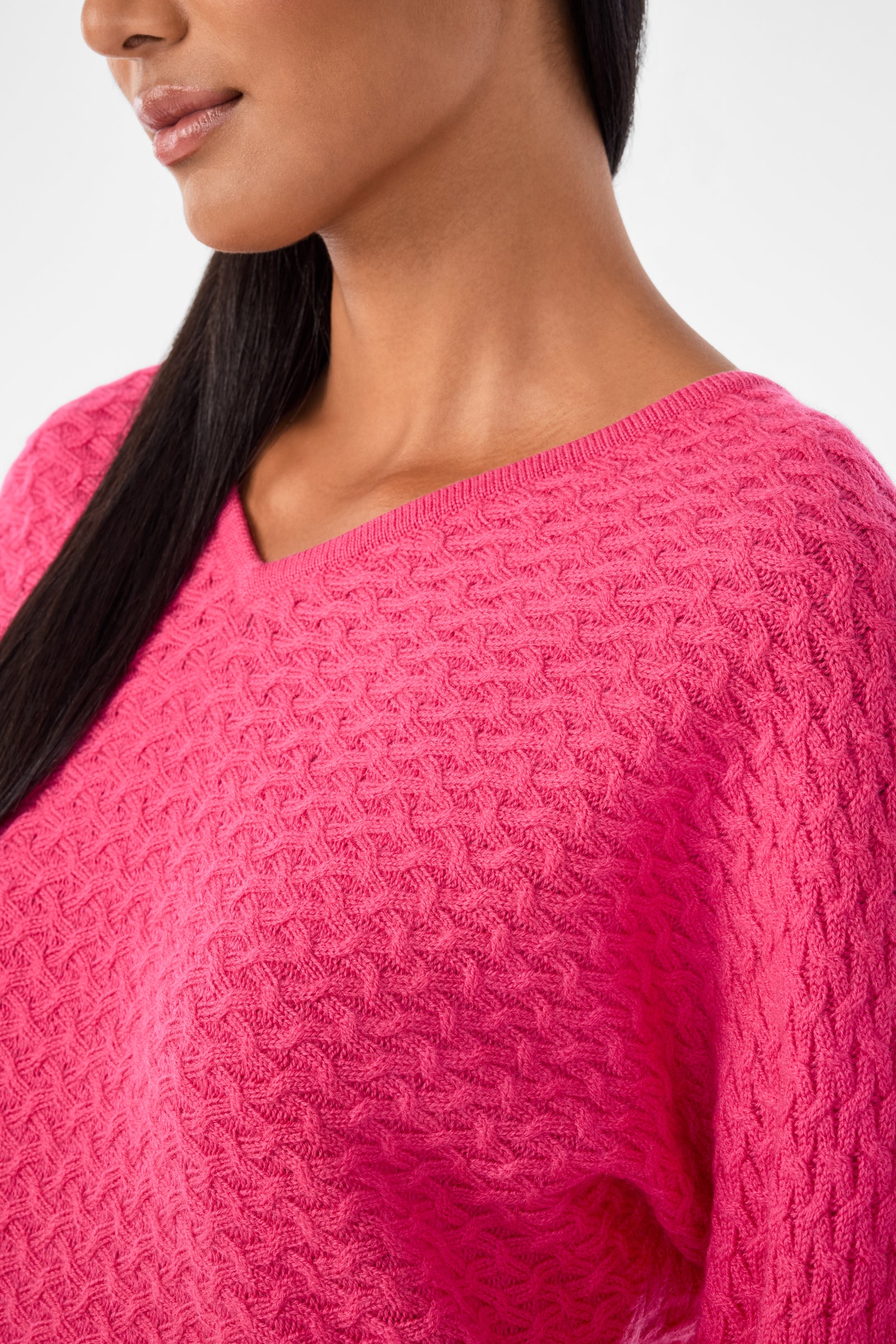 A close-up of a woman wearing the Lima V-Neck Pullover, a textured bright pink sweater. Only the lower half of her face, neck, shoulder, and part of her chest are shown, with straight dark hair draped over one shoulder.