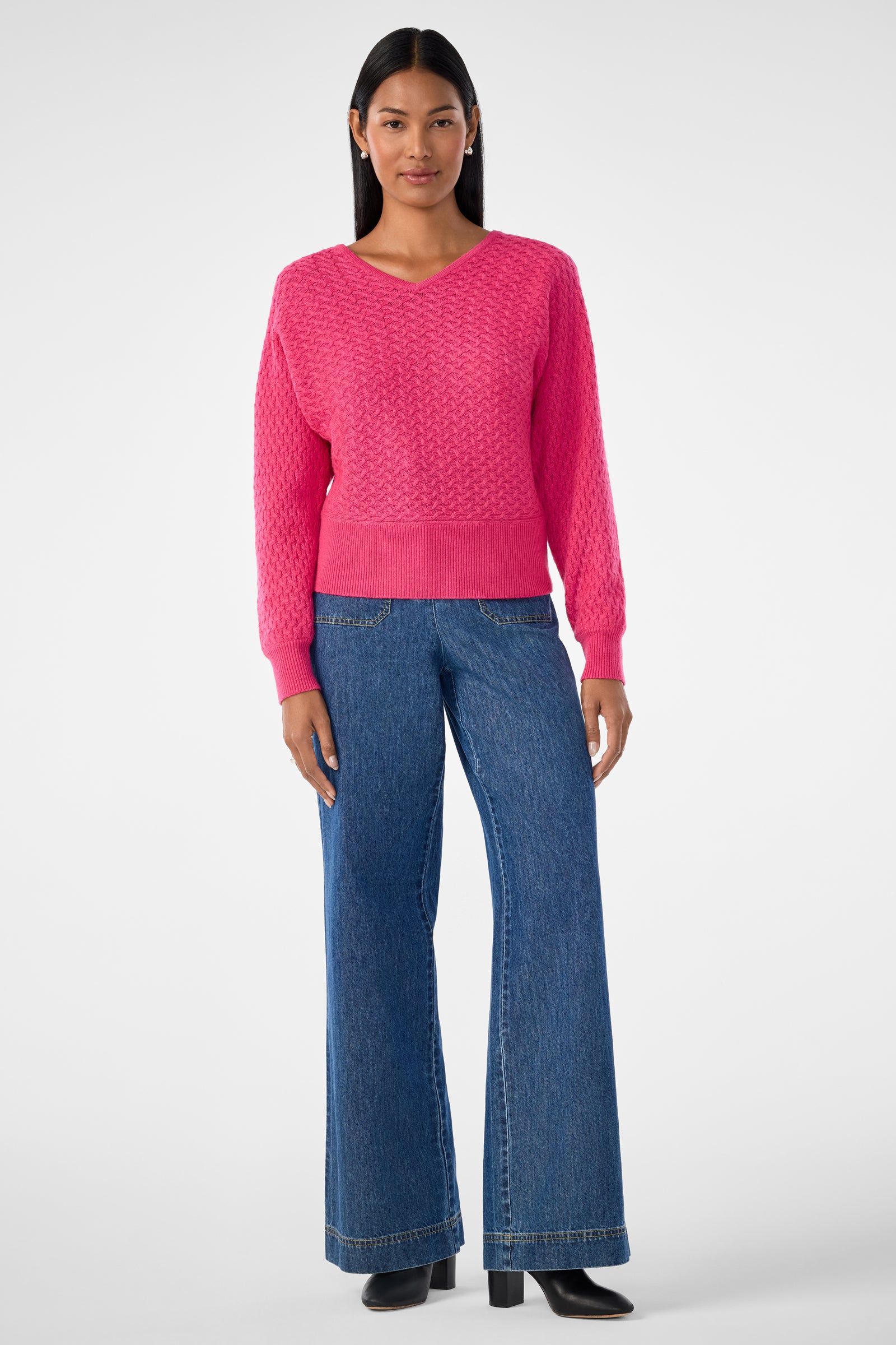 A woman stands facing forward, wearing the Lima V-Neck Pullover in bright pink and wide-leg blue jeans with black heeled boots against a plain, light background.