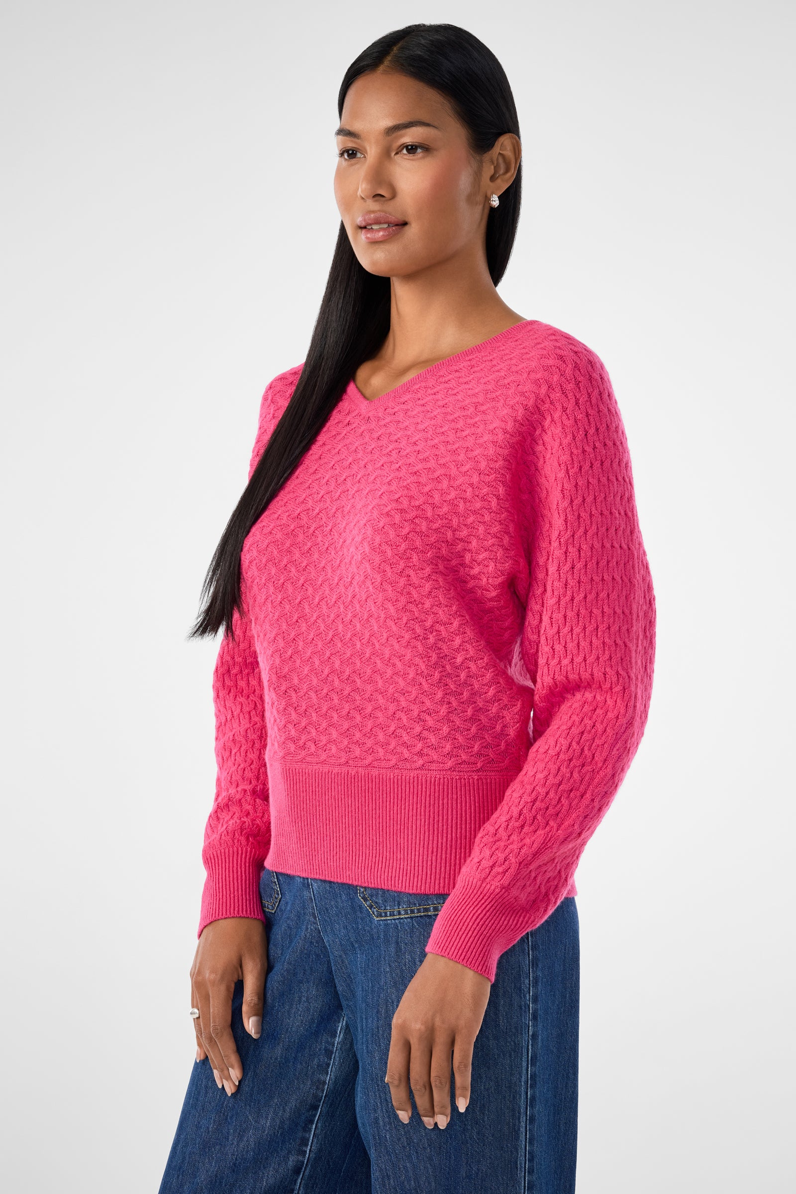 A woman with long black hair wears the Lima V-Neck Pullover in bright pink and blue jeans, standing against a light grey background and looking slightly to the side with a neutral expression.