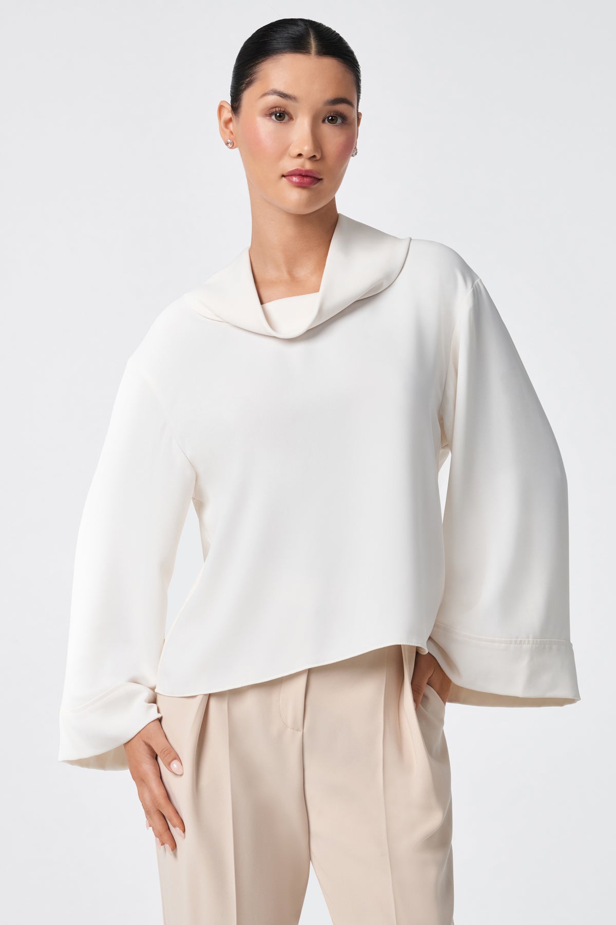 A woman with dark hair in a sleek bun wears the Staged Top, a white, long-sleeve blouse with a wide collar and loose fit, paired with beige pleated pants. She poses with one hand in her pocket against a plain light background.