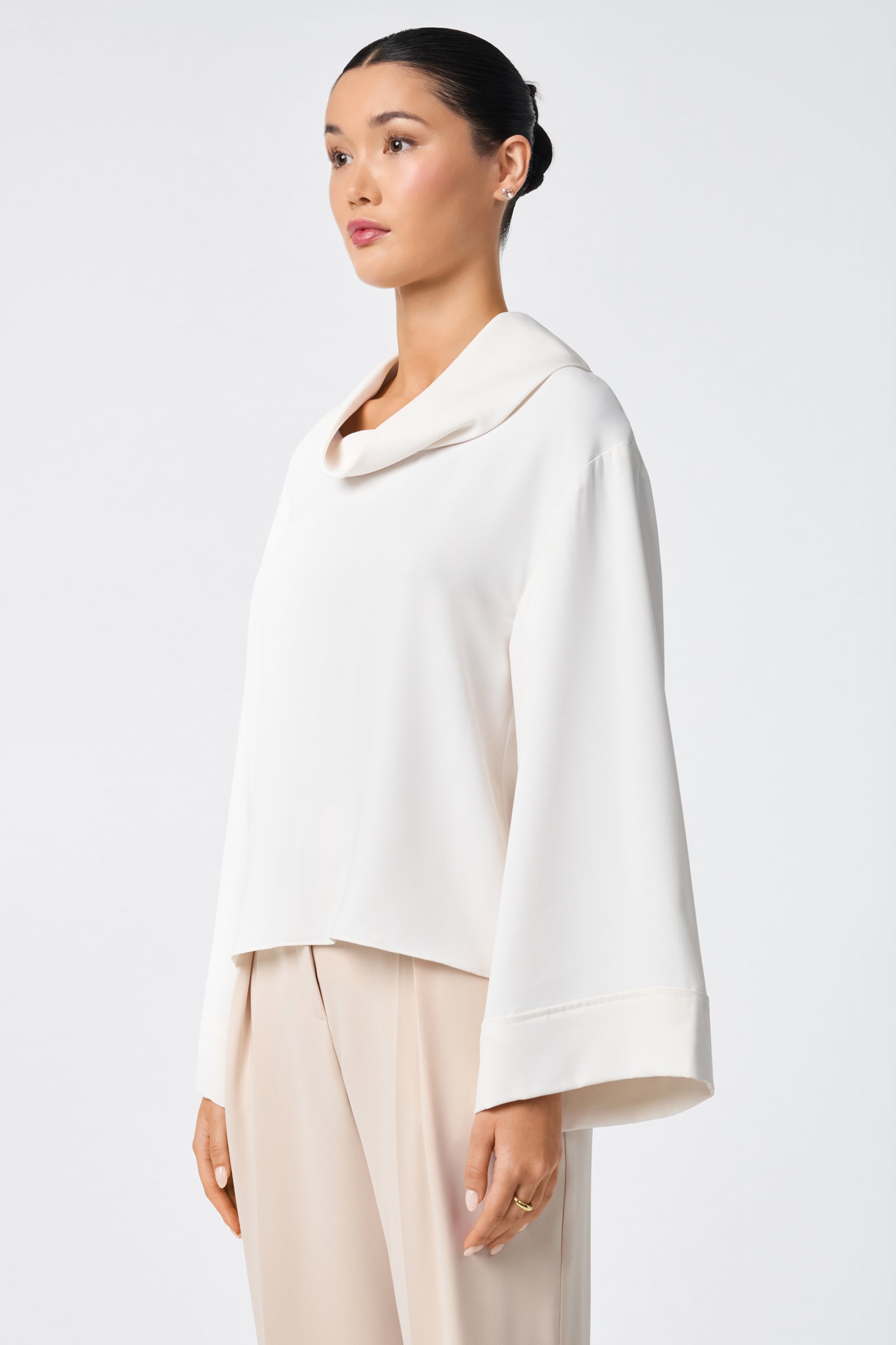 A woman in profile stands against a white background, wearing the Staged Top—a loose-fitting, long-sleeve cream blouse with a wide collar—paired with matching wide-leg pants. Her hair is pulled back neatly, and she looks calm.