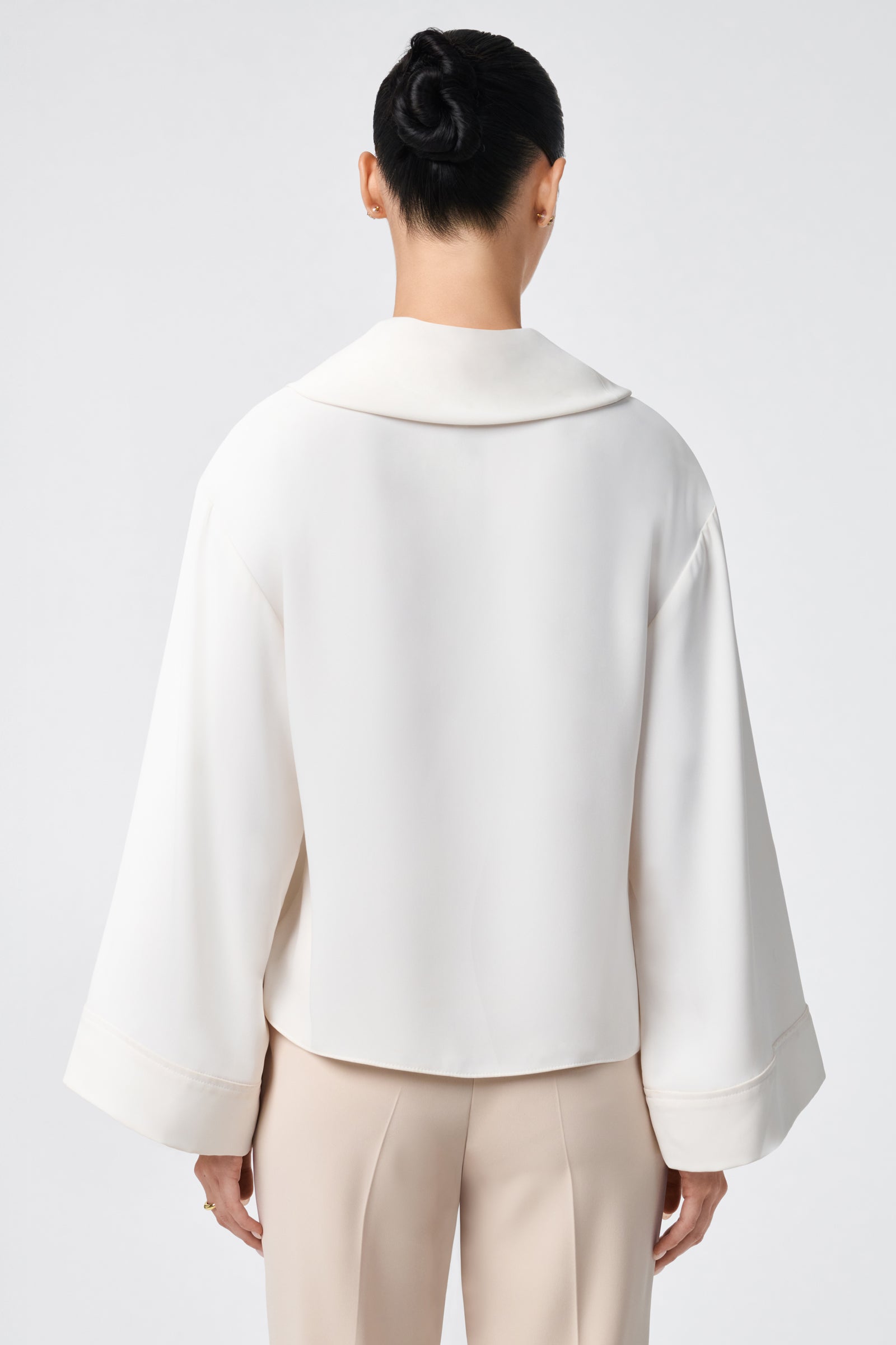 A person with dark hair in a neat bun is seen from behind, wearing the loose-fitting ivory Staged Top with wide sleeves and beige pants, set against a plain light background.