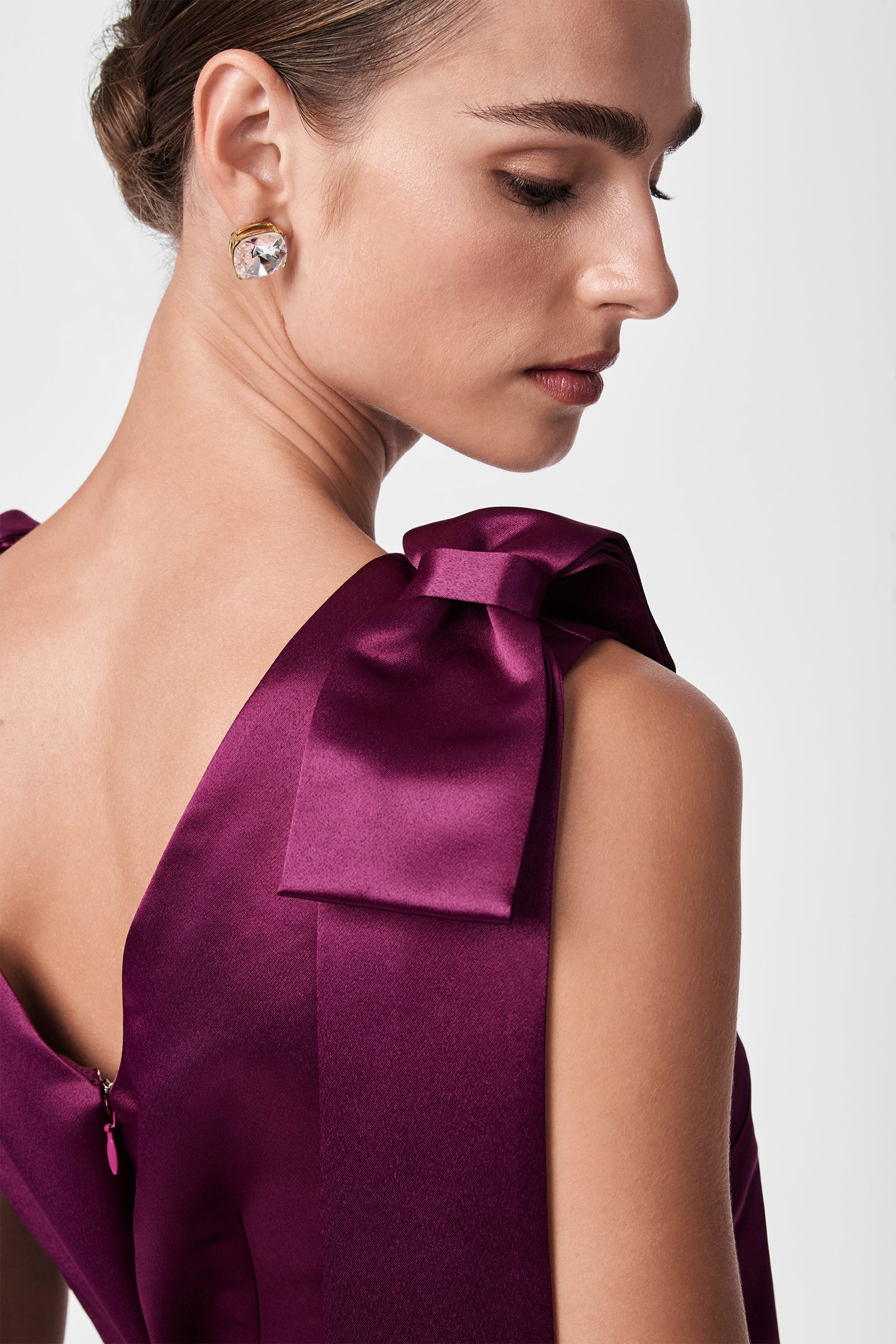A woman with brown hair in an updo wears the Priscilla Dress, a deep purple satin piece featuring large shoulder bows and a visible back zipper. She accessorizes with round gold earrings and gazes down over her shoulder.