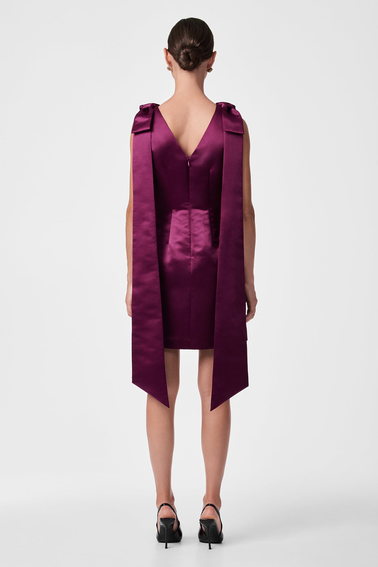 A woman faces away, wearing the Priscilla Dress— a sleeveless, deep purple satin mini dress with long, wide shoulder ribbons. Her hair is neatly tied back, and she pairs the look with black heeled shoes against a plain white background.