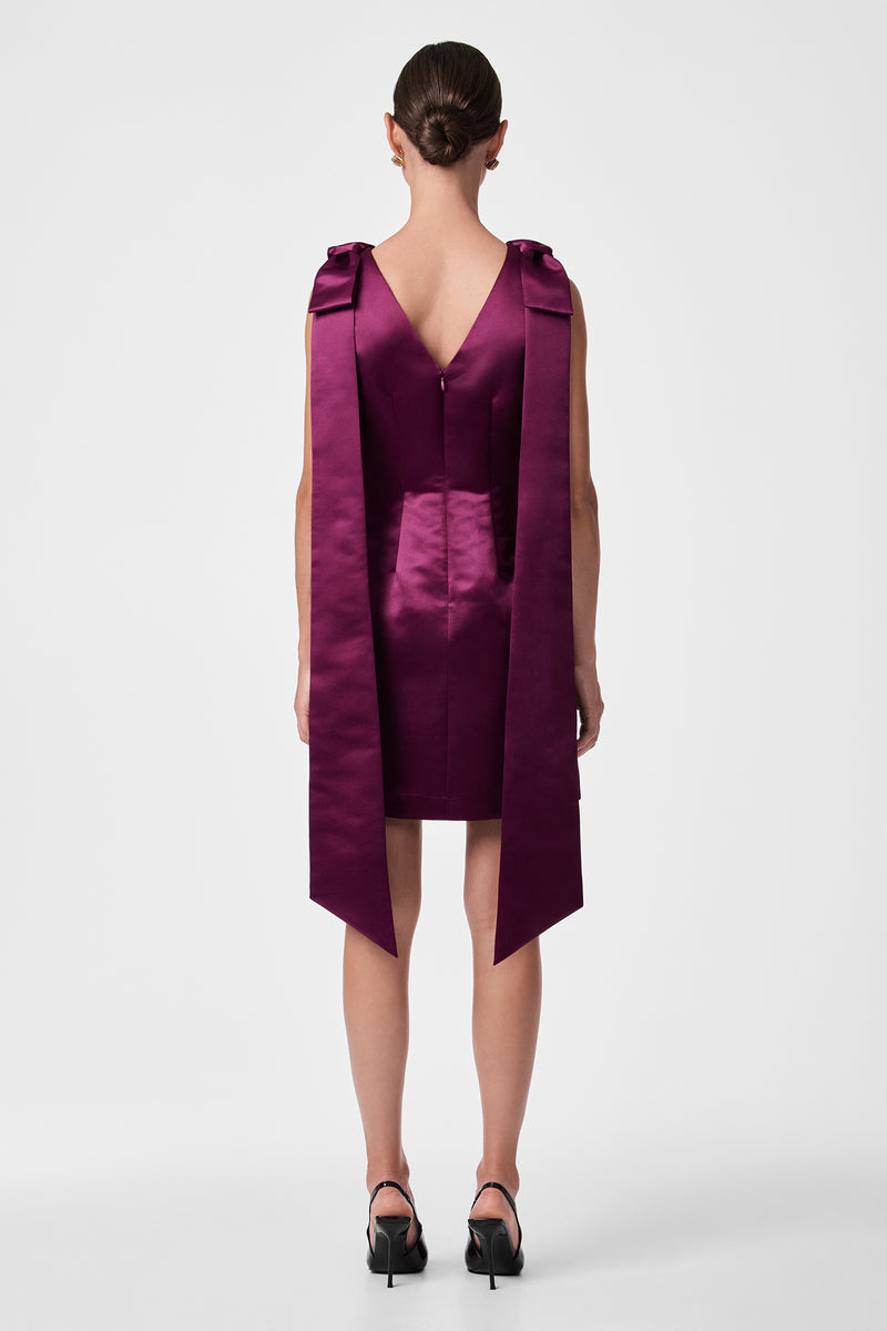 A woman faces away, wearing the Priscilla Dress— a sleeveless, deep purple satin mini dress with long, wide shoulder ribbons. Her hair is neatly tied back, and she pairs the look with black heeled shoes against a plain white background.