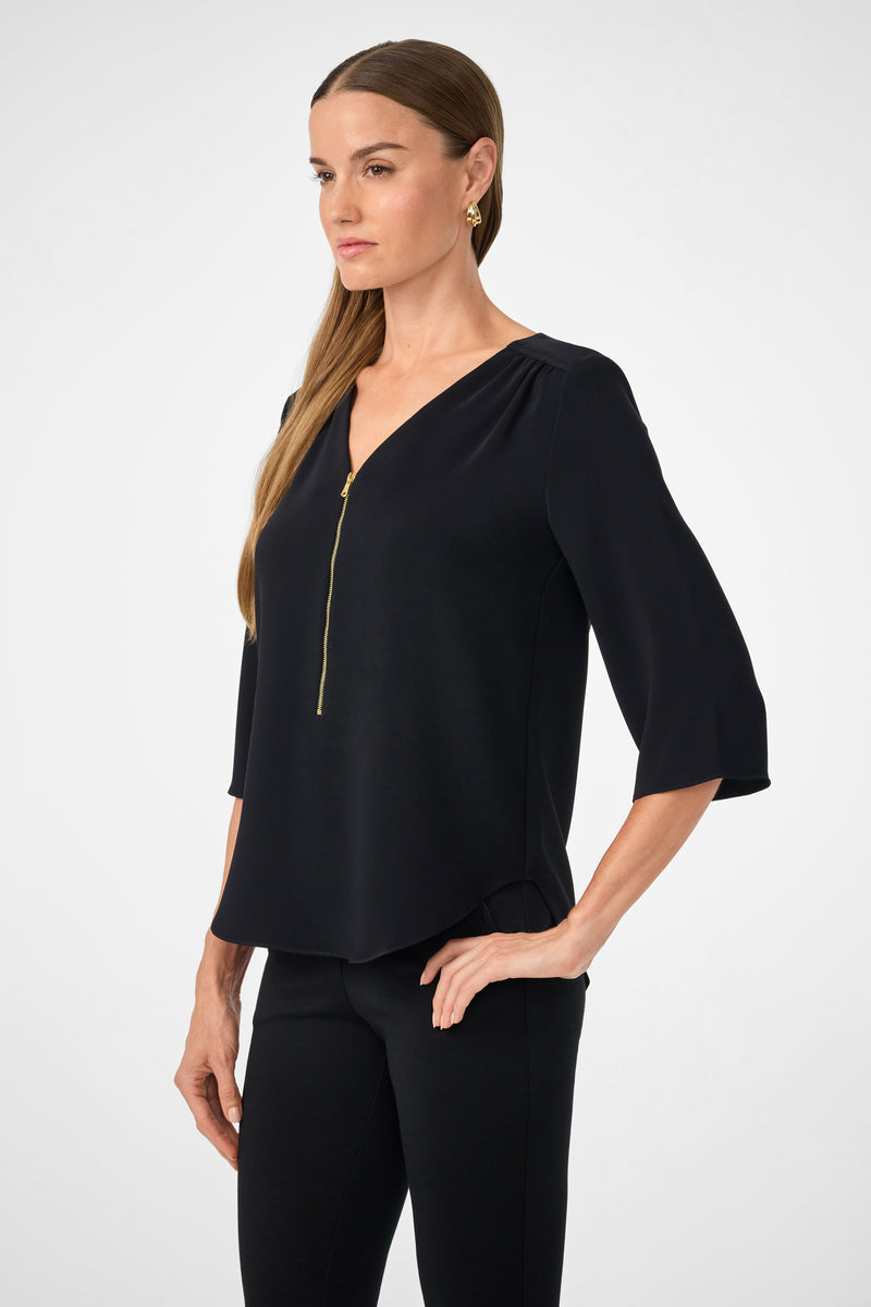 A woman with long brown hair models the Tamarac Top, a black blouse with three-quarter sleeves and a V-neck zip detail, standing with one hand on her hip against a plain white background.